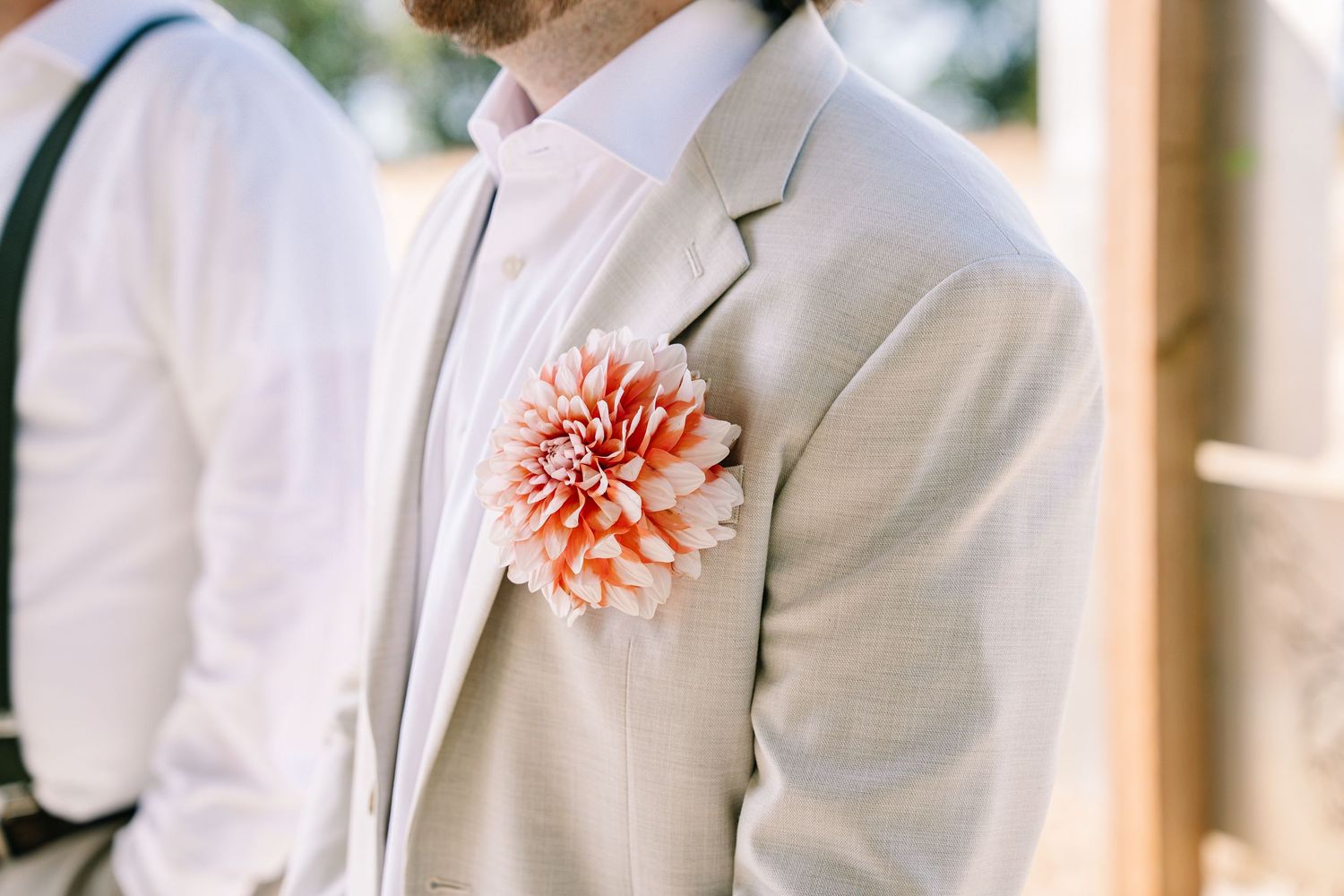 A row of groomsmen wearing white tuxedo jackets with coral boutonnieres stand in front of wooden columns.