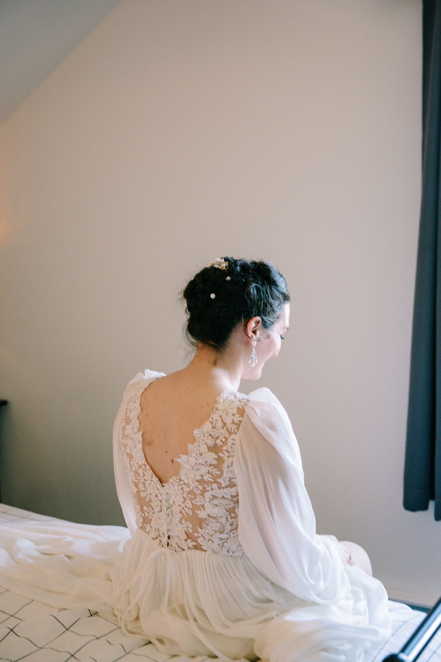 A person in a flowing white lace dress with long sleeves sits elegantly on a bed in a minimalist room.