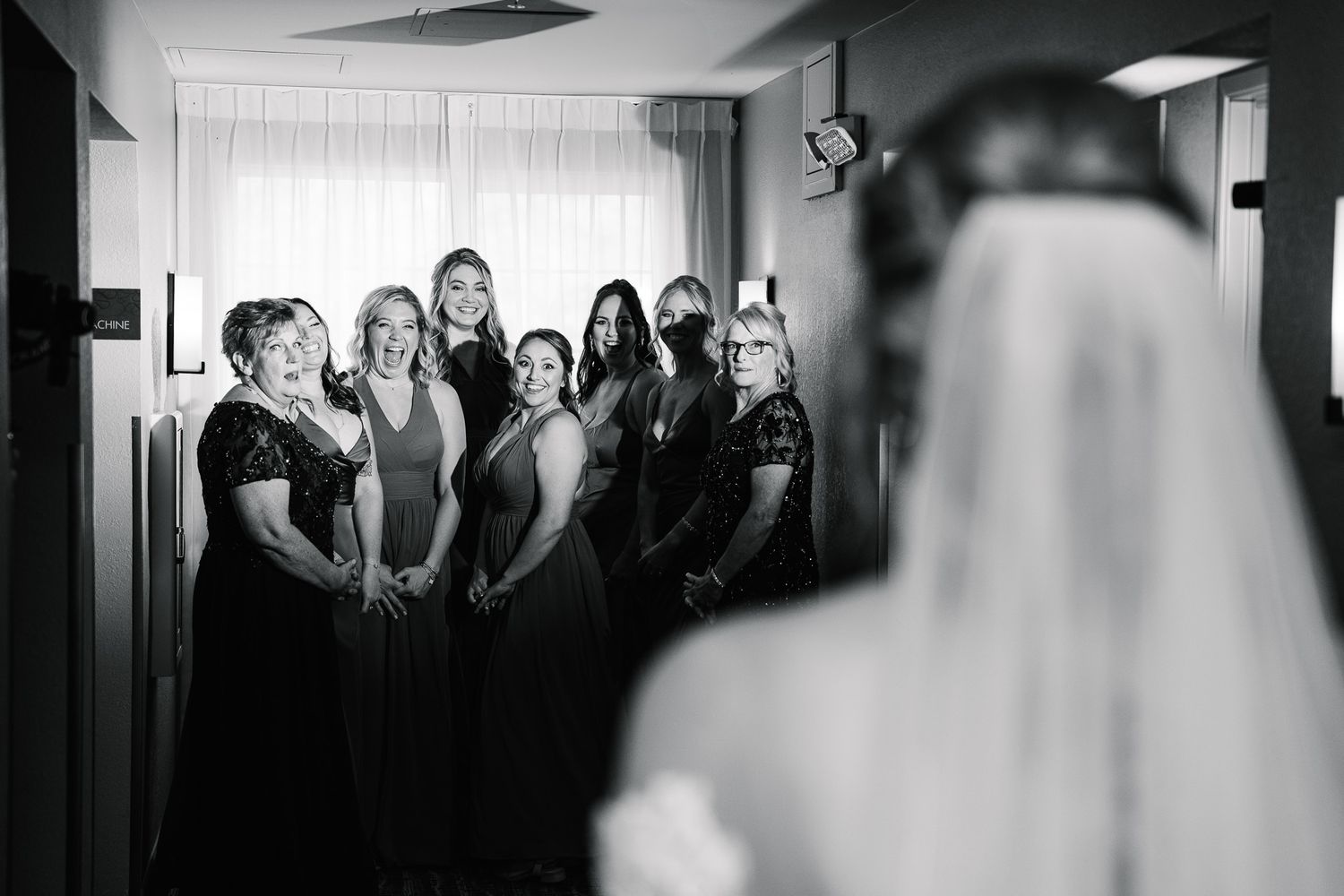 Black and white candid wedding photo showing a group in matching dresses reacting together in a hallway moment.