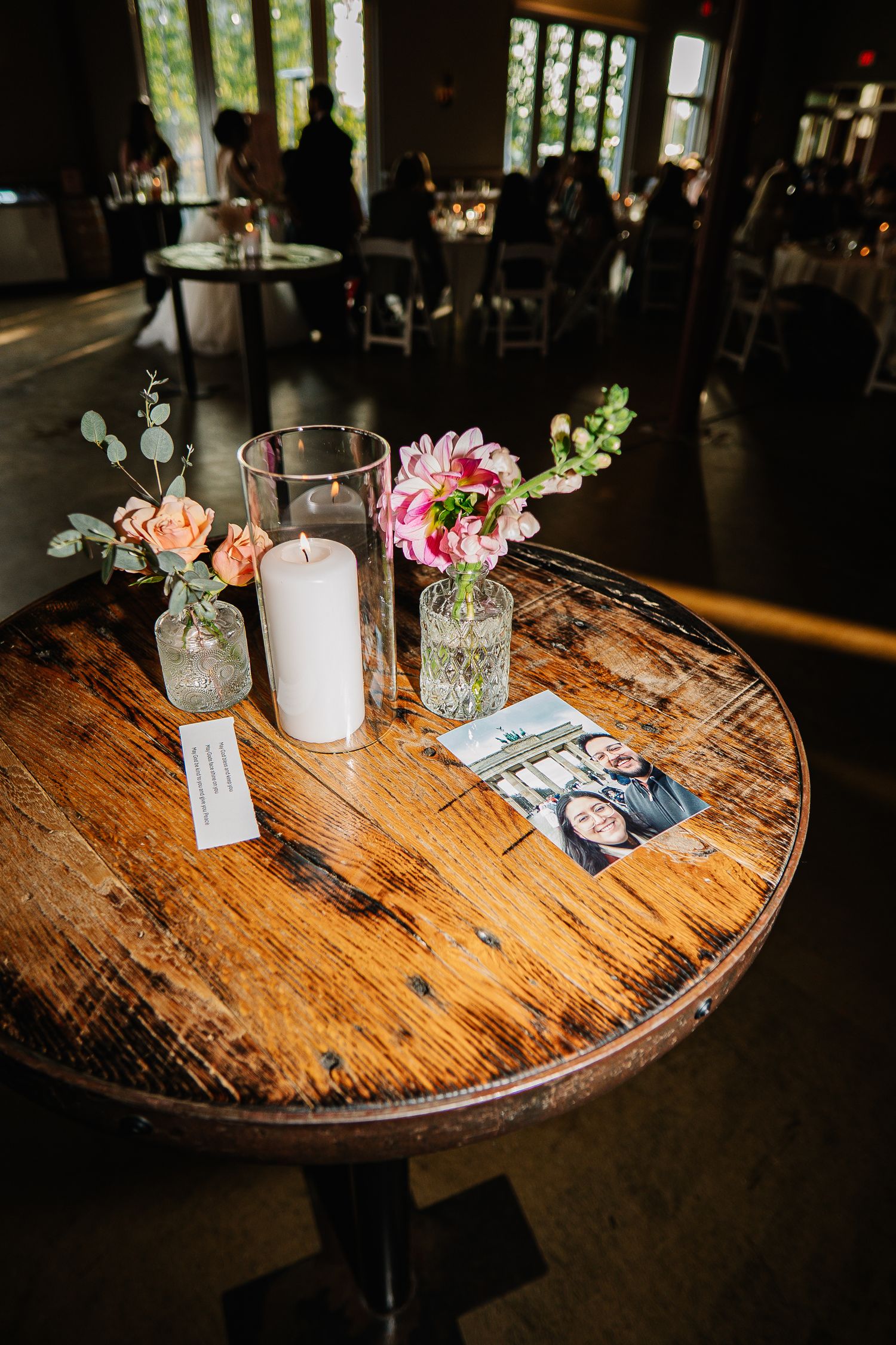 Rustic wooden round tables with small glass vases holding delicate floral arrangements and candles in a dimly lit venue.