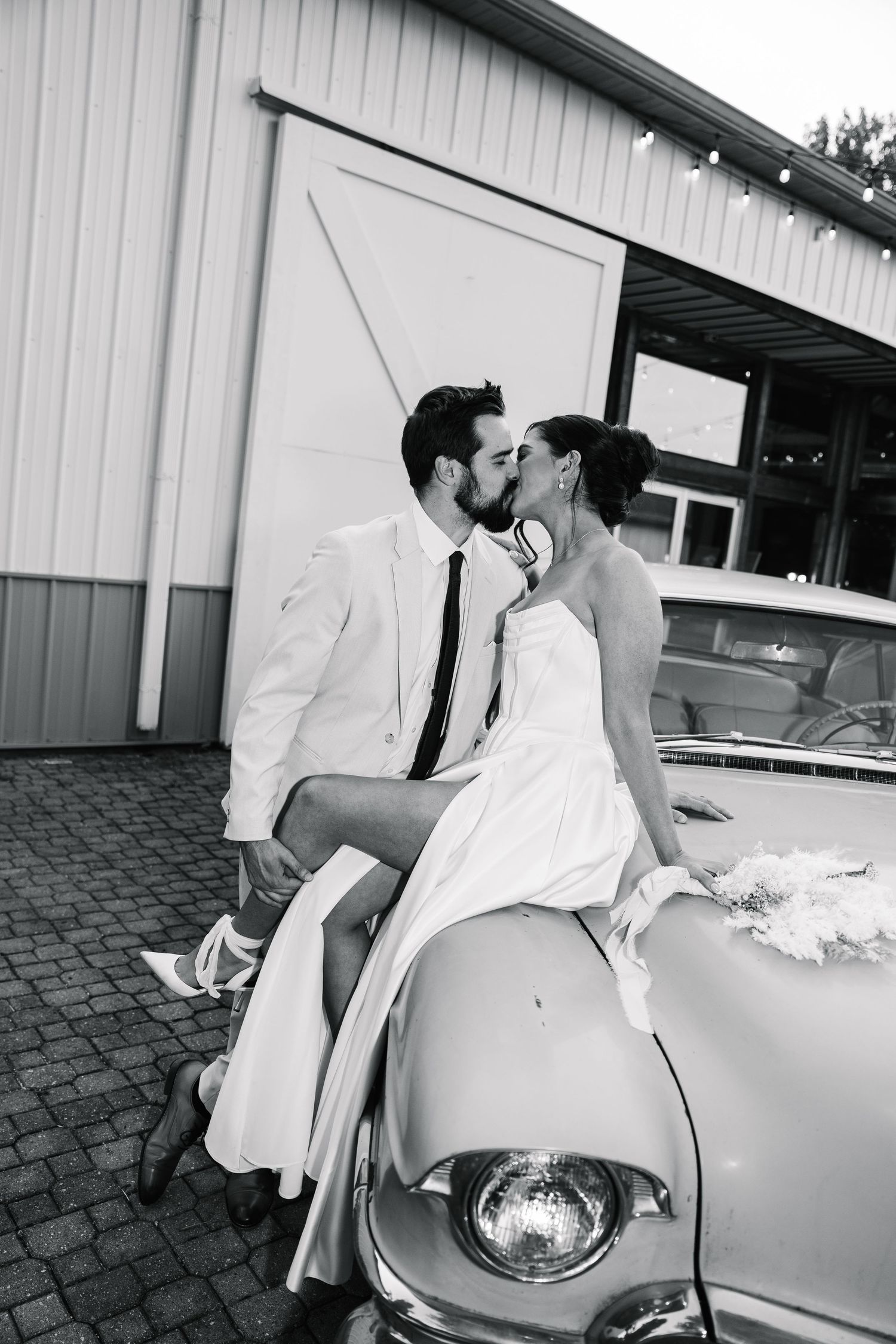 Black and white photo of a romantic couple in wedding attire posing on a vintage car outside a building.