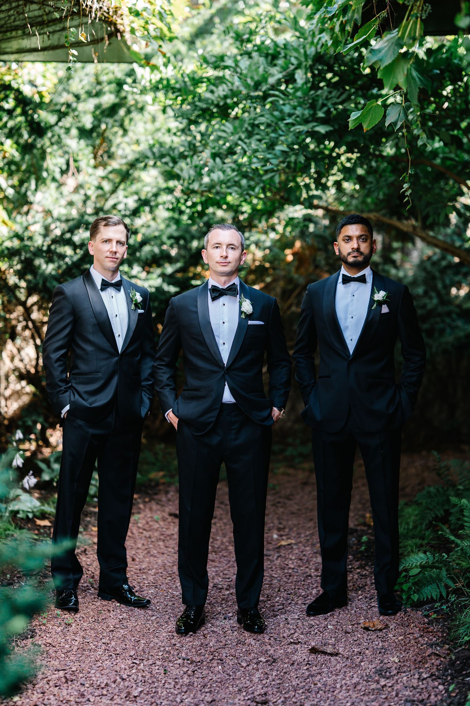 Groomsmen in classic black tuxedos pose together on a wooded path during a wedding photoshoot.