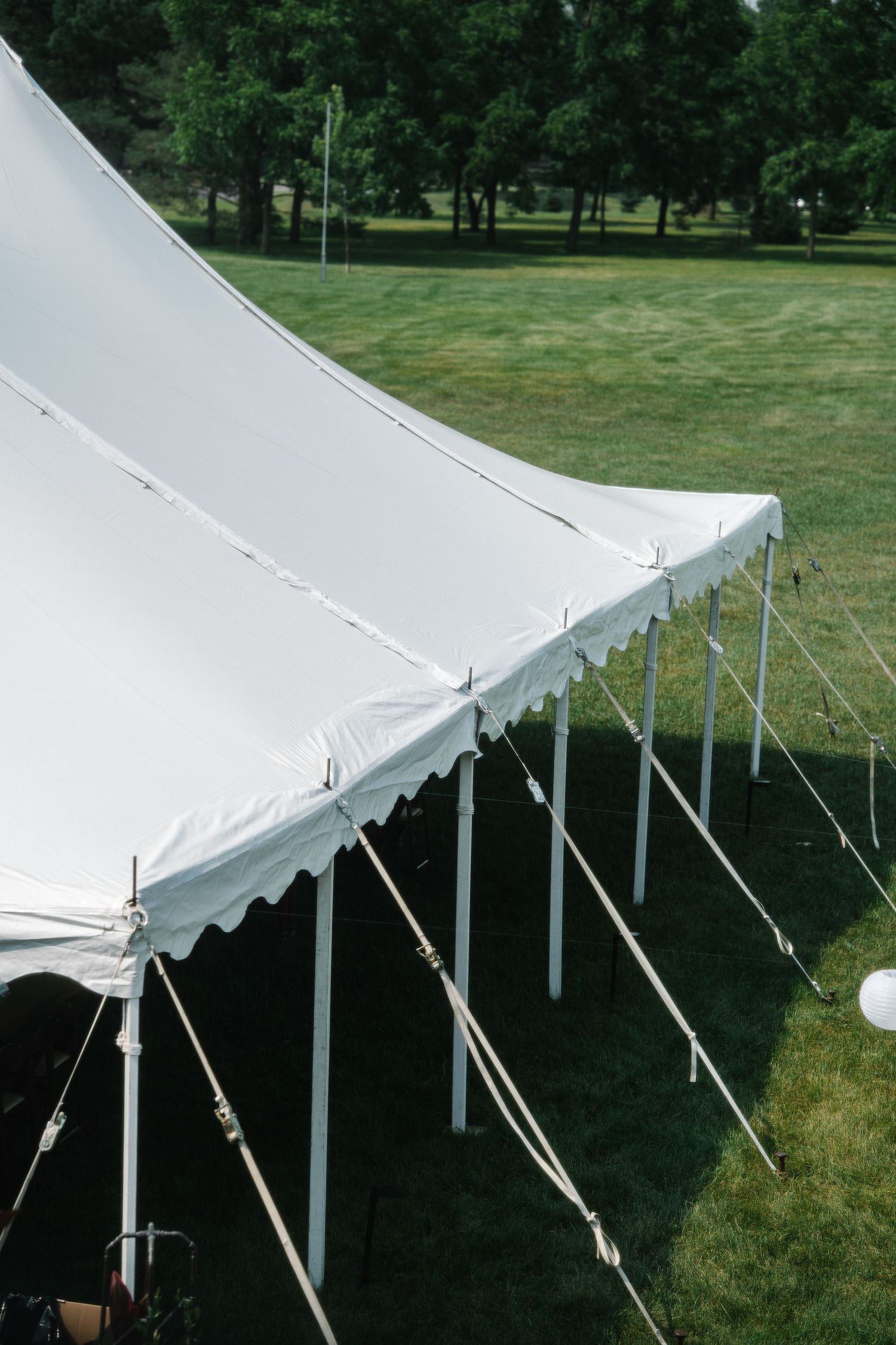 White outdoor event tent with poles and supports casting shadows on green grass in a park setting.