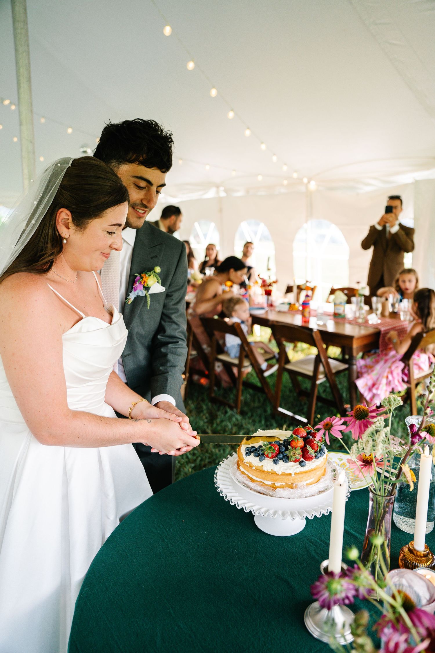 Couple cuts their wedding cake decorated with fresh berries on a white cake stand under an elegant reception tent.