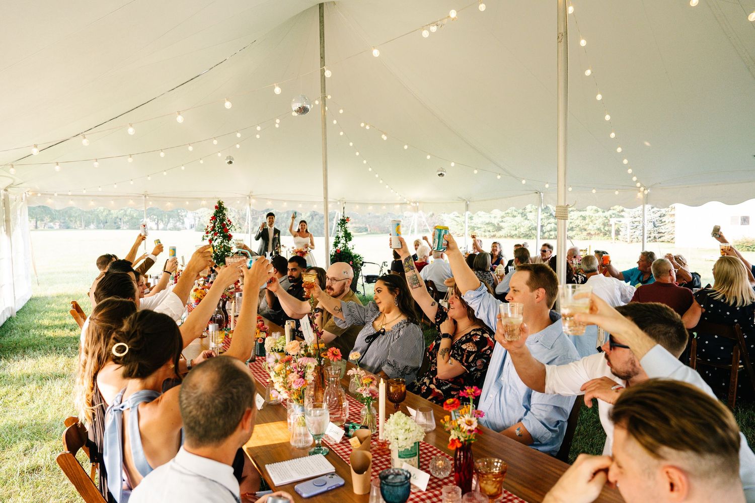 Wedding guests raise their glasses in a toast under a white tent decorated with string lights during an outdoor celebration.