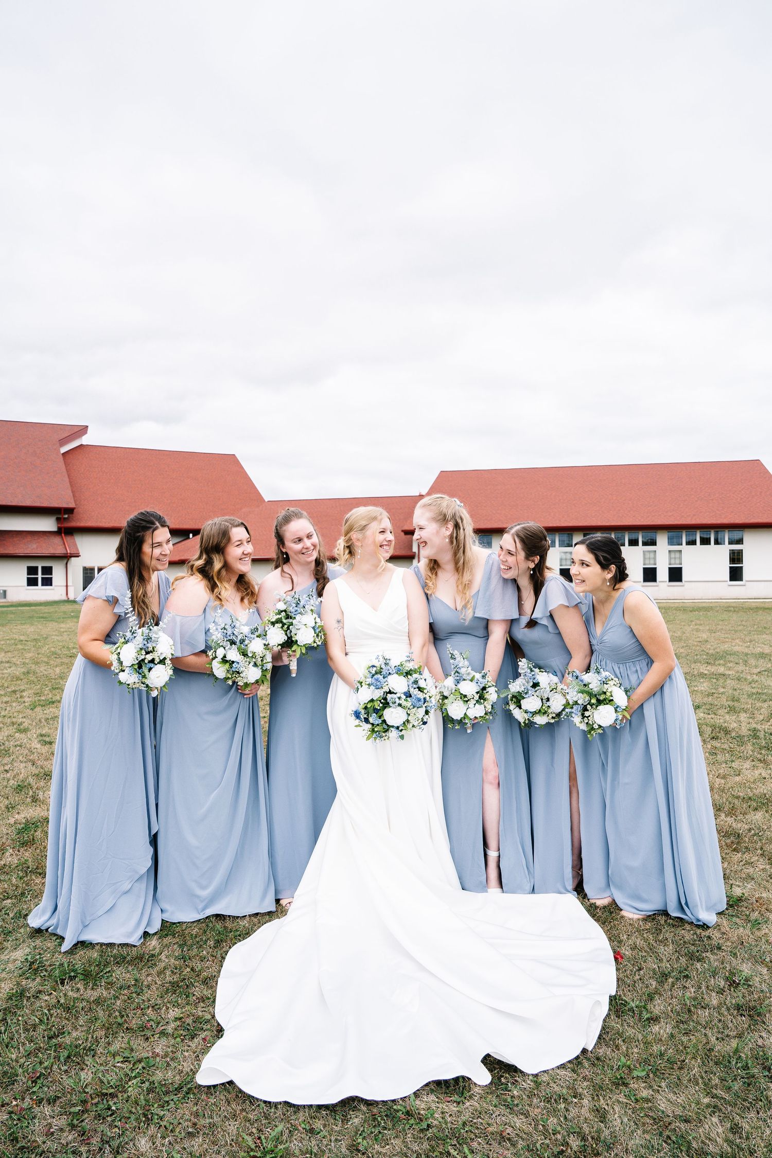 A bridal party in light blue bridesmaid dresses and white wedding gown pose together on grass outside a building.