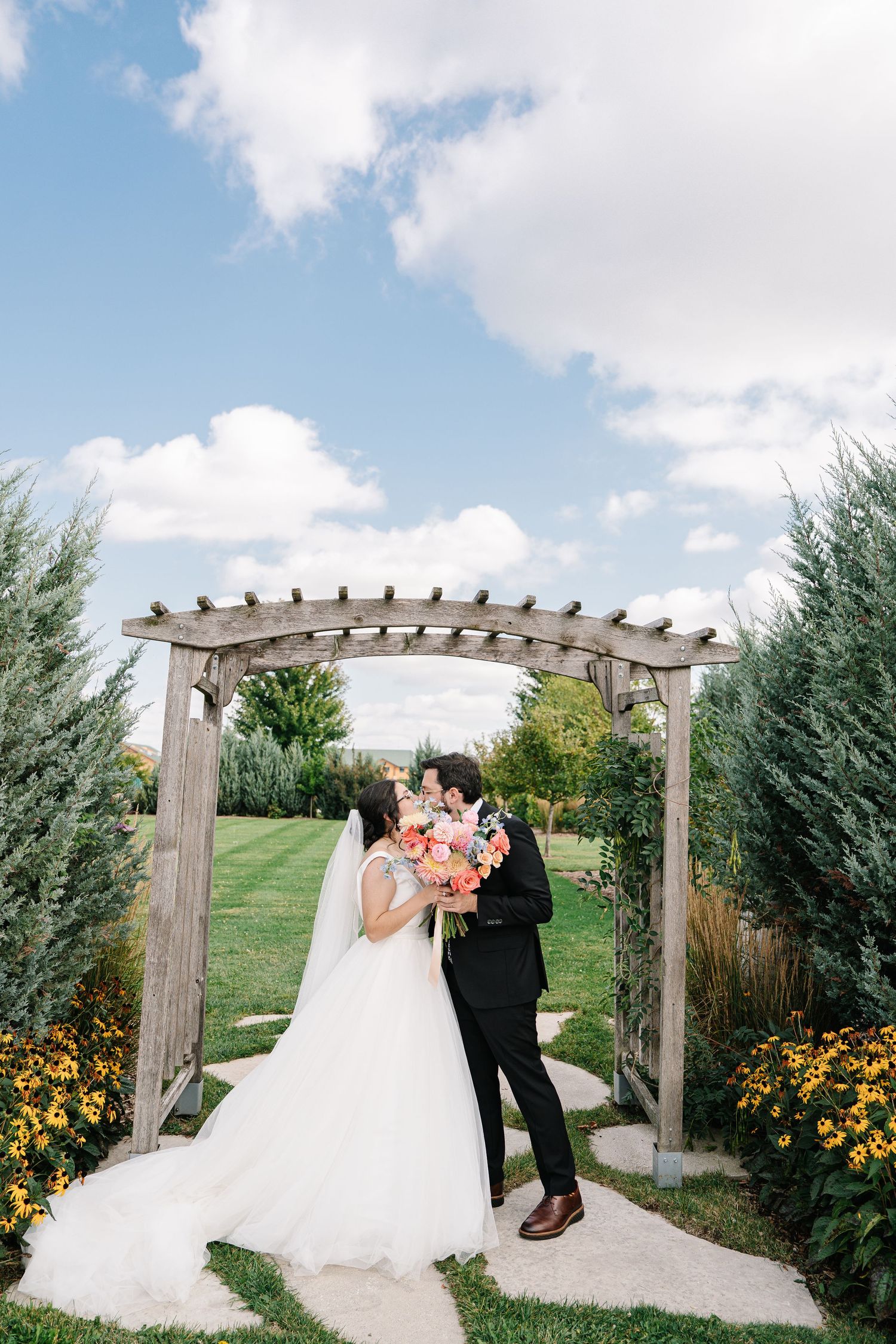 Newlyweds share a romantic moment under a wooden pergola surrounded by yellow flowers and greenery.