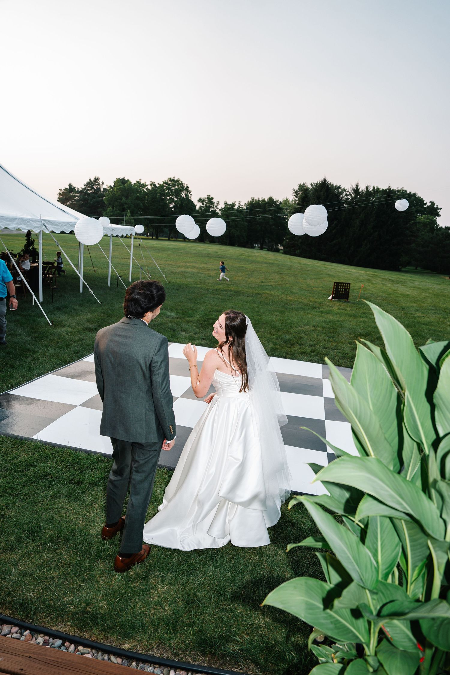 A couple in wedding attire walks through a green garden setting with white paper lanterns and decorative plants.
