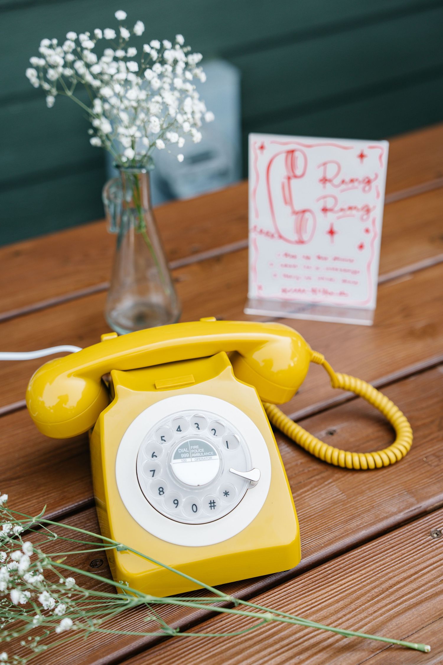 A vintage yellow rotary telephone with white baby's breath flowers sits on a wooden table in a decorative setting.