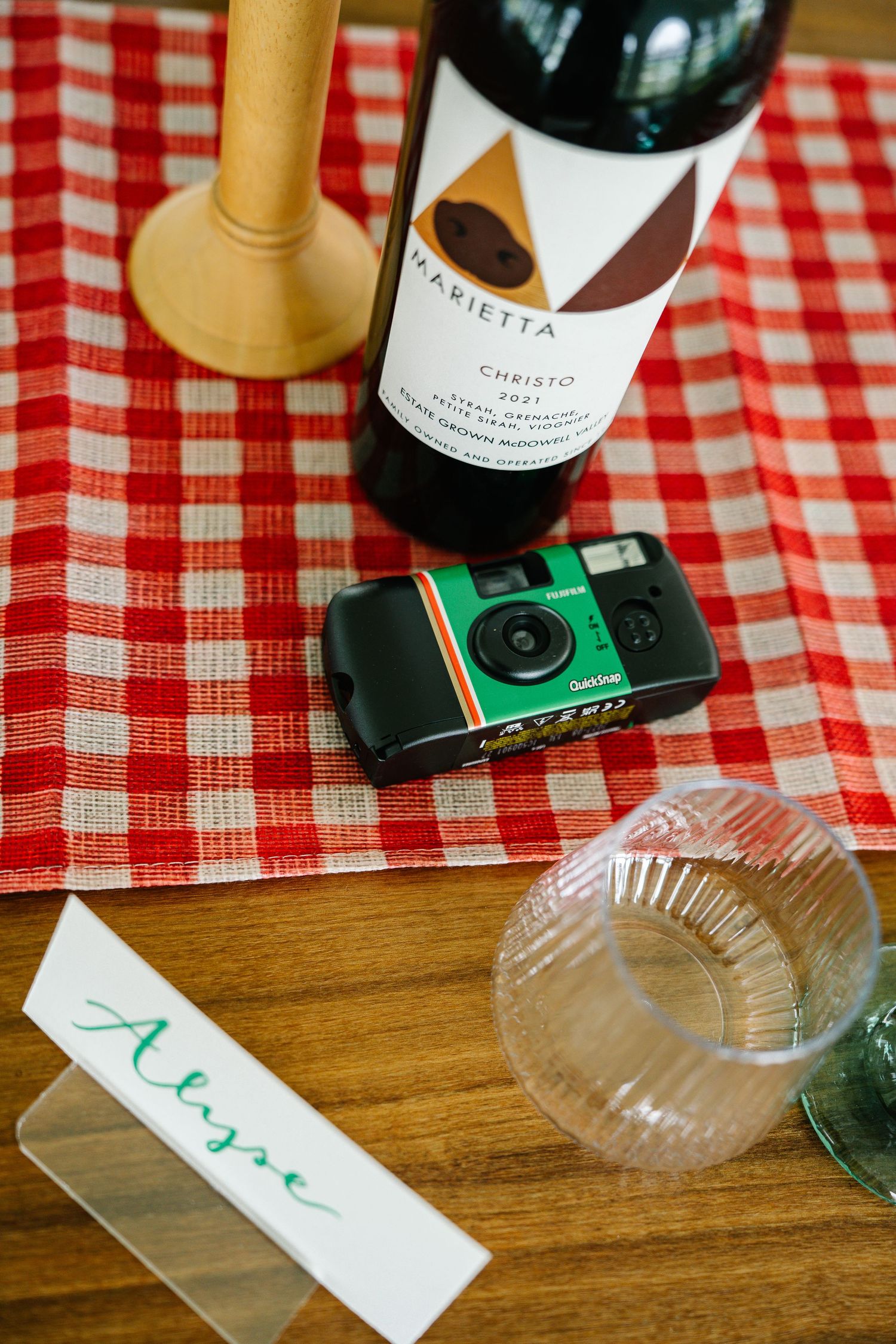 Wine bottles and disposable cameras arranged on a red and white checkered tablecloth at a party setting.