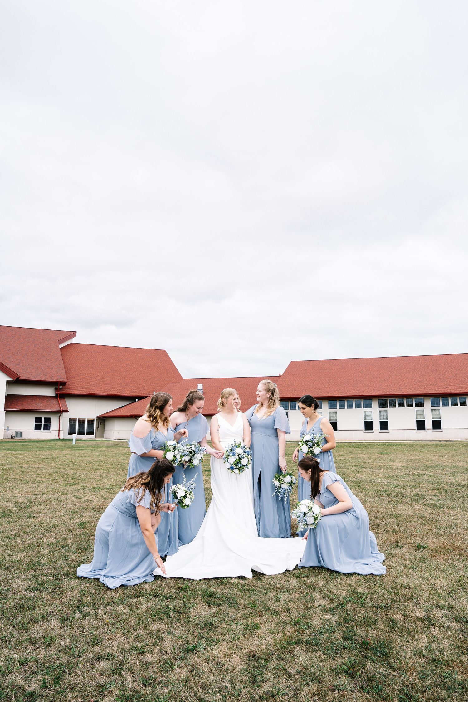 Wedding party wearing light blue dresses and white gown pose together on grassy field near red barn building.