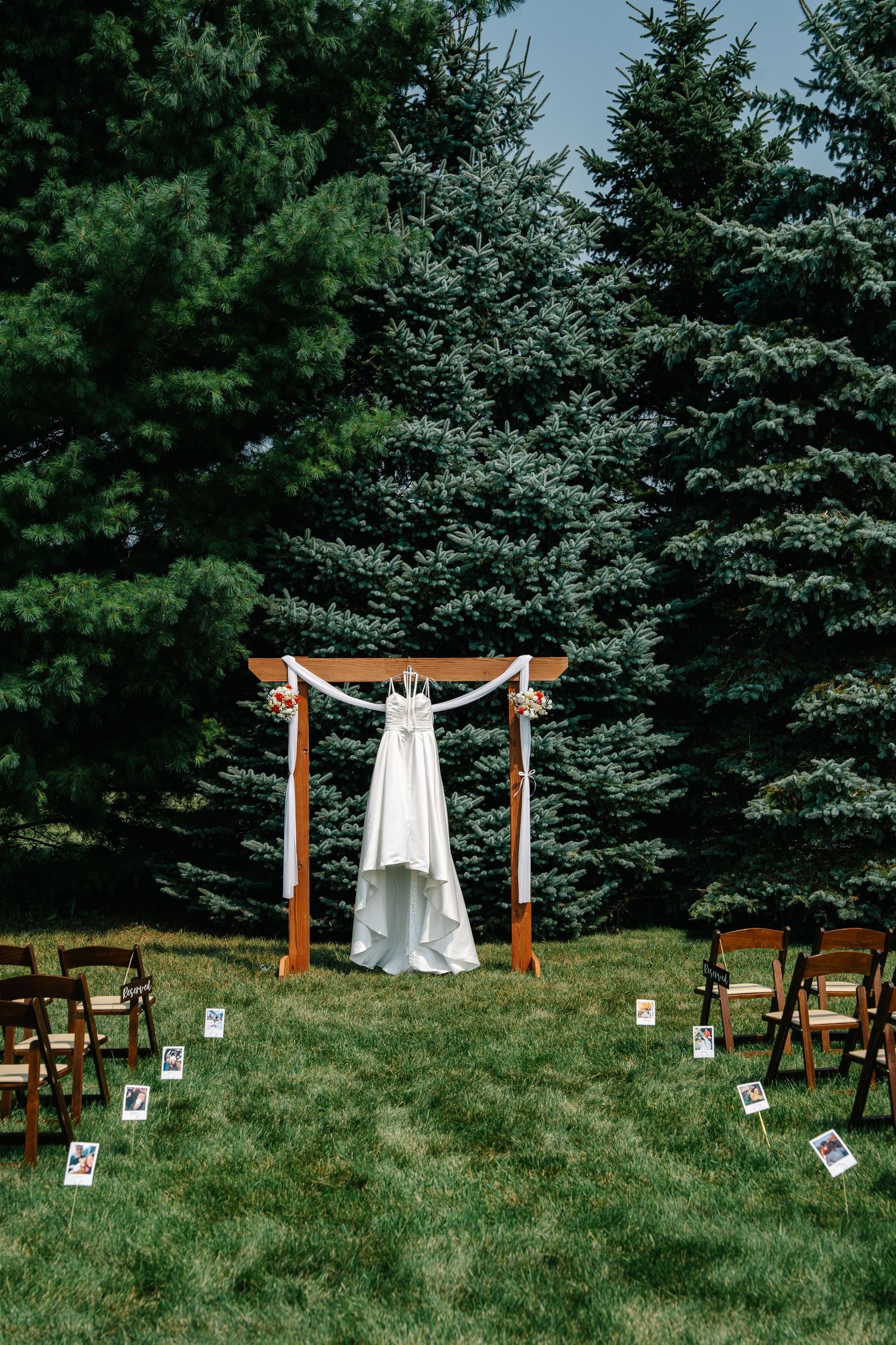White wedding dress hangs elegantly from wooden arch in outdoor forest ceremony setting with scattered chairs on grass.