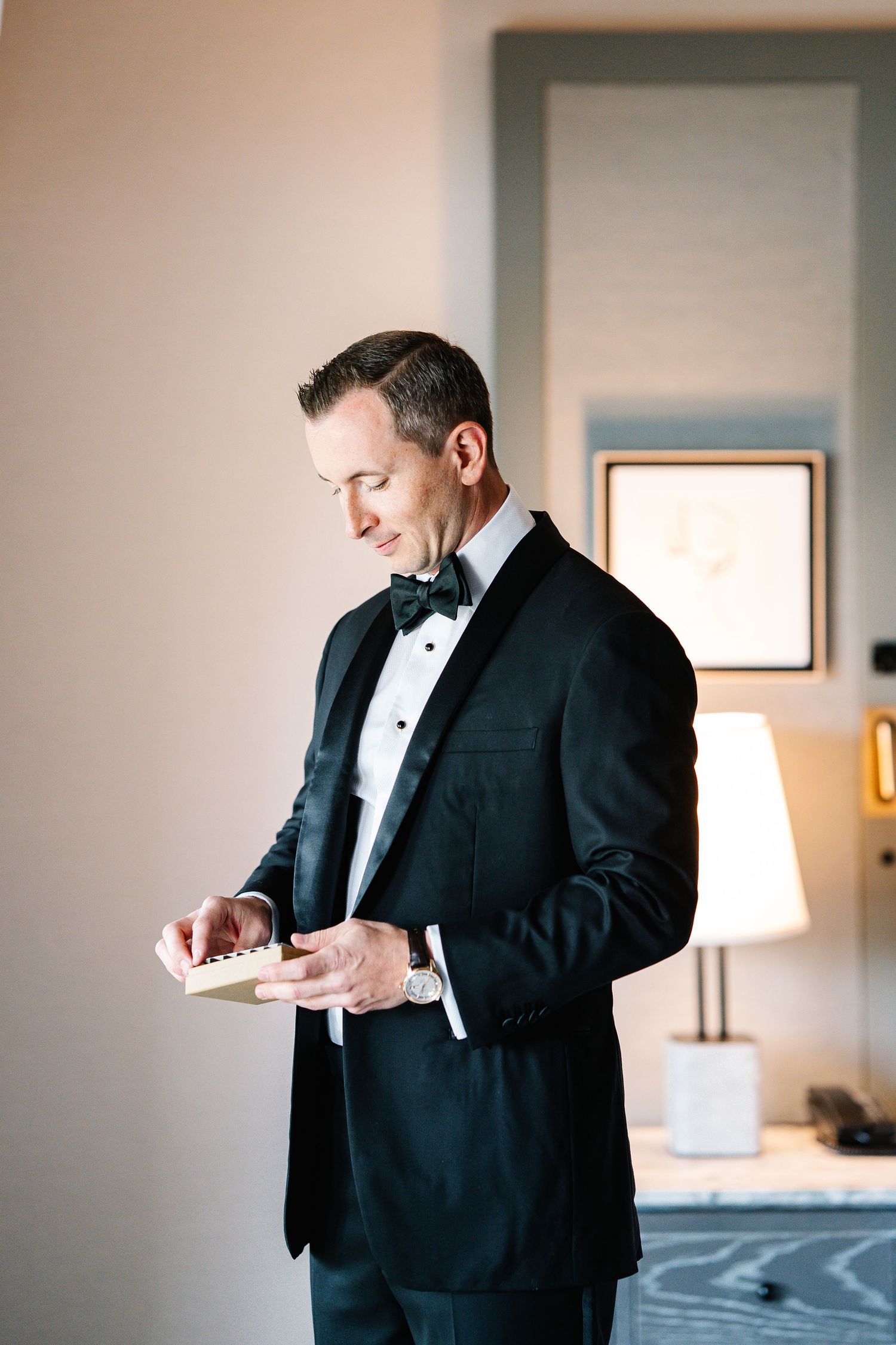 A groom in a classic black tuxedo and bow tie adjusts his cuffs while getting ready in a bright hotel room.