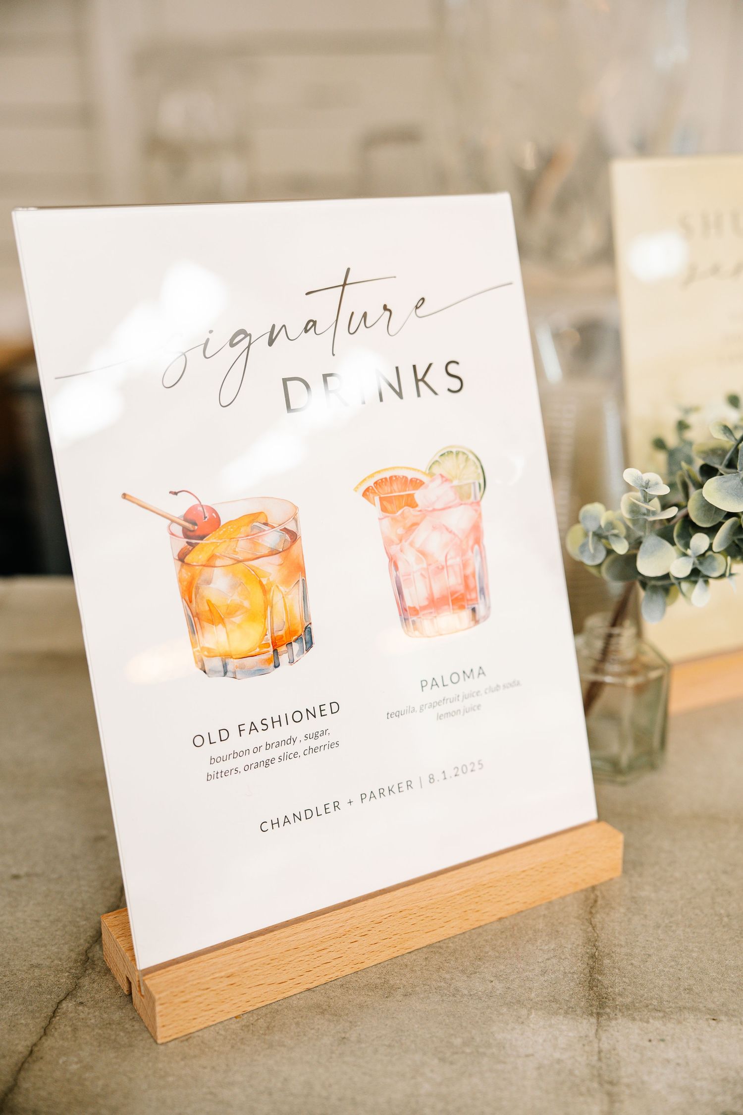 Elegant wedding signature drinks menu featuring watercolor illustrations of an Old Fashioned and Paloma cocktail in a wooden stand.