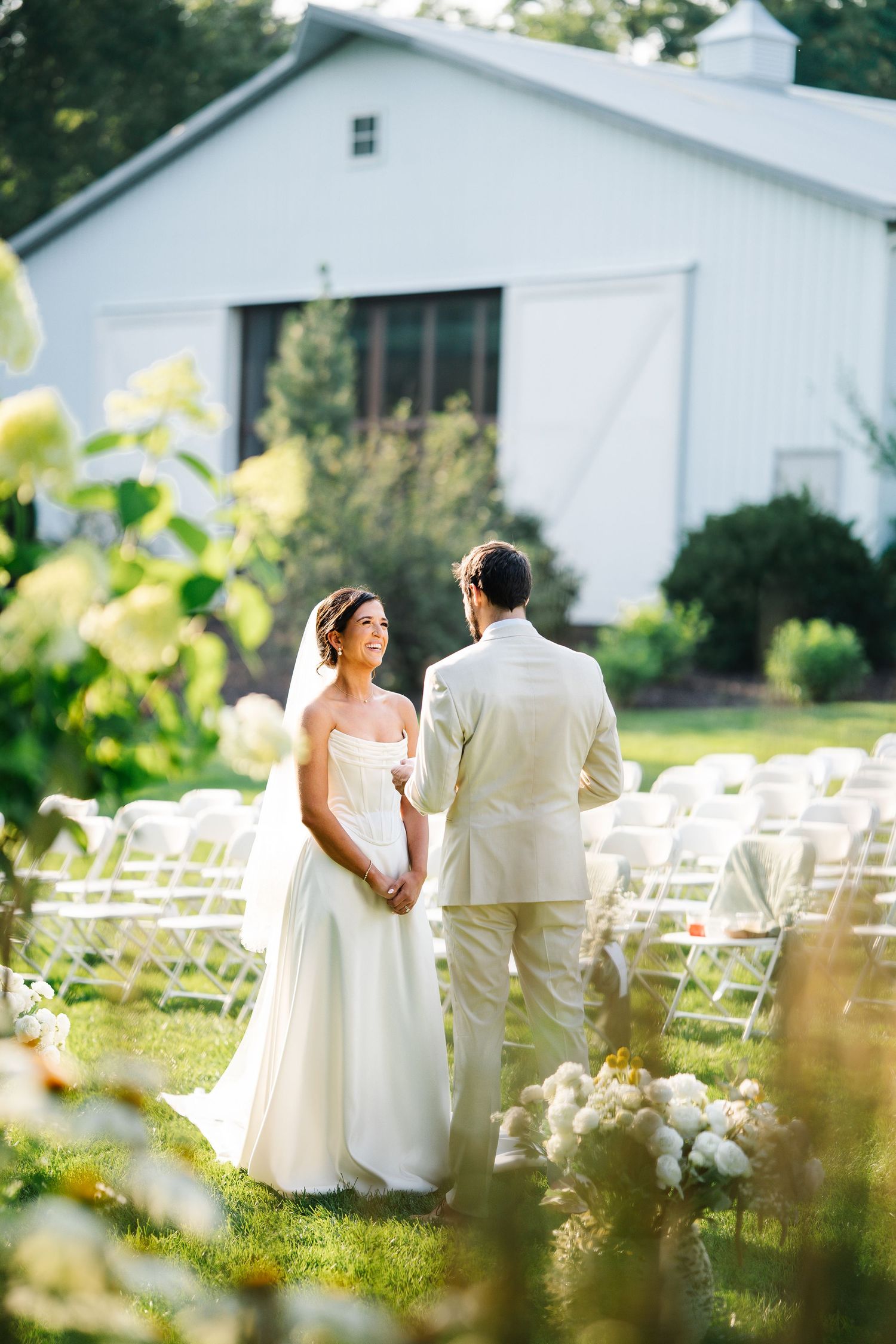 A romantic outdoor wedding ceremony setup with white chairs and a rustic barn backdrop in soft natural lighting.