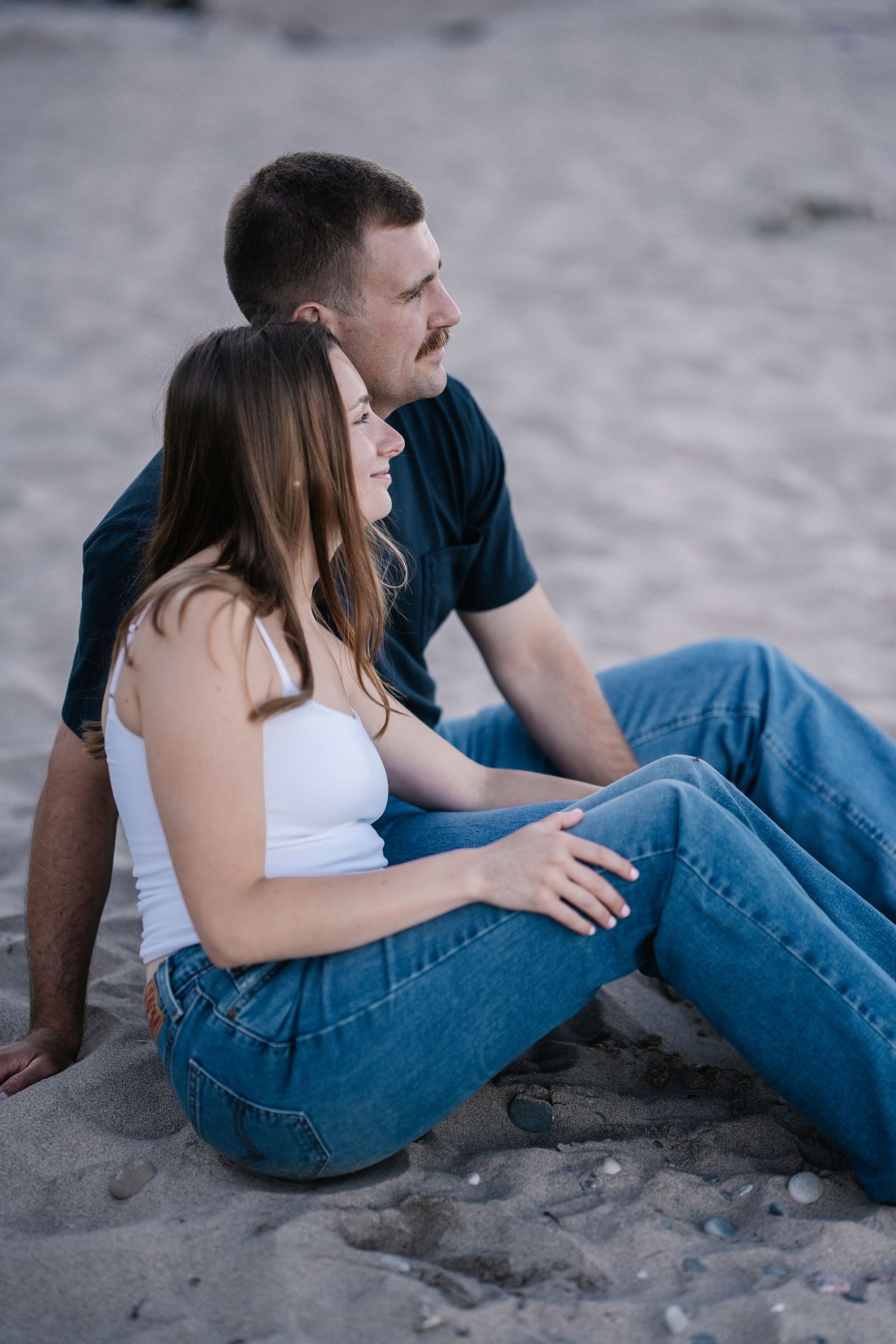 Couple sitting together on sandy beach, woman in white top and jeans leaning against man in navy shirt.