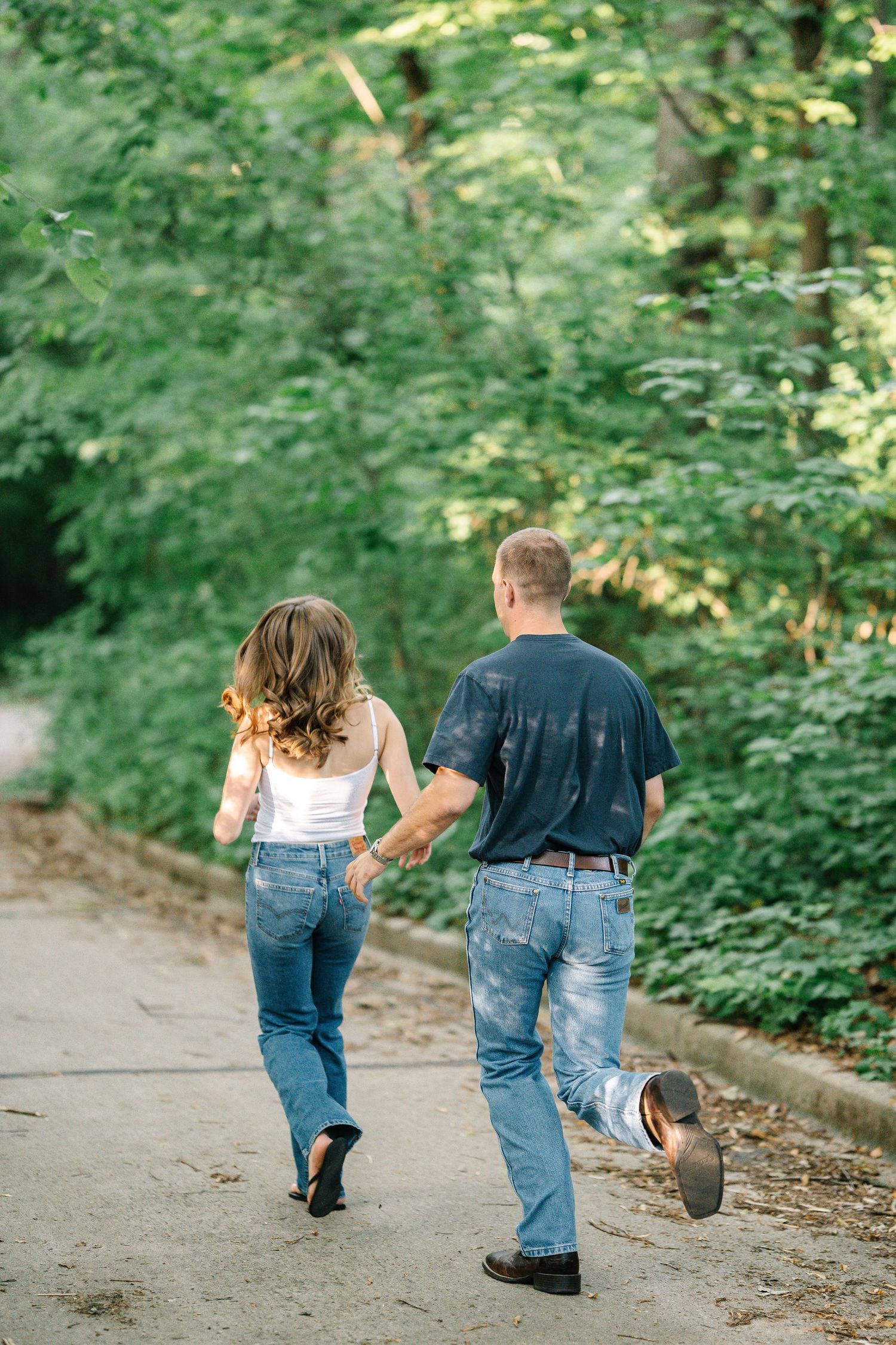 Couple holding hands walking down a tree-lined path, wearing jeans, viewed from behind.