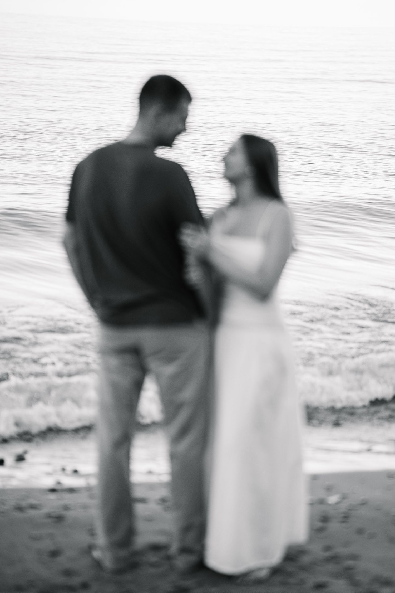 Blurry black and white photo of a couple holding hands on a beach, facing each other with ocean waves behind them.