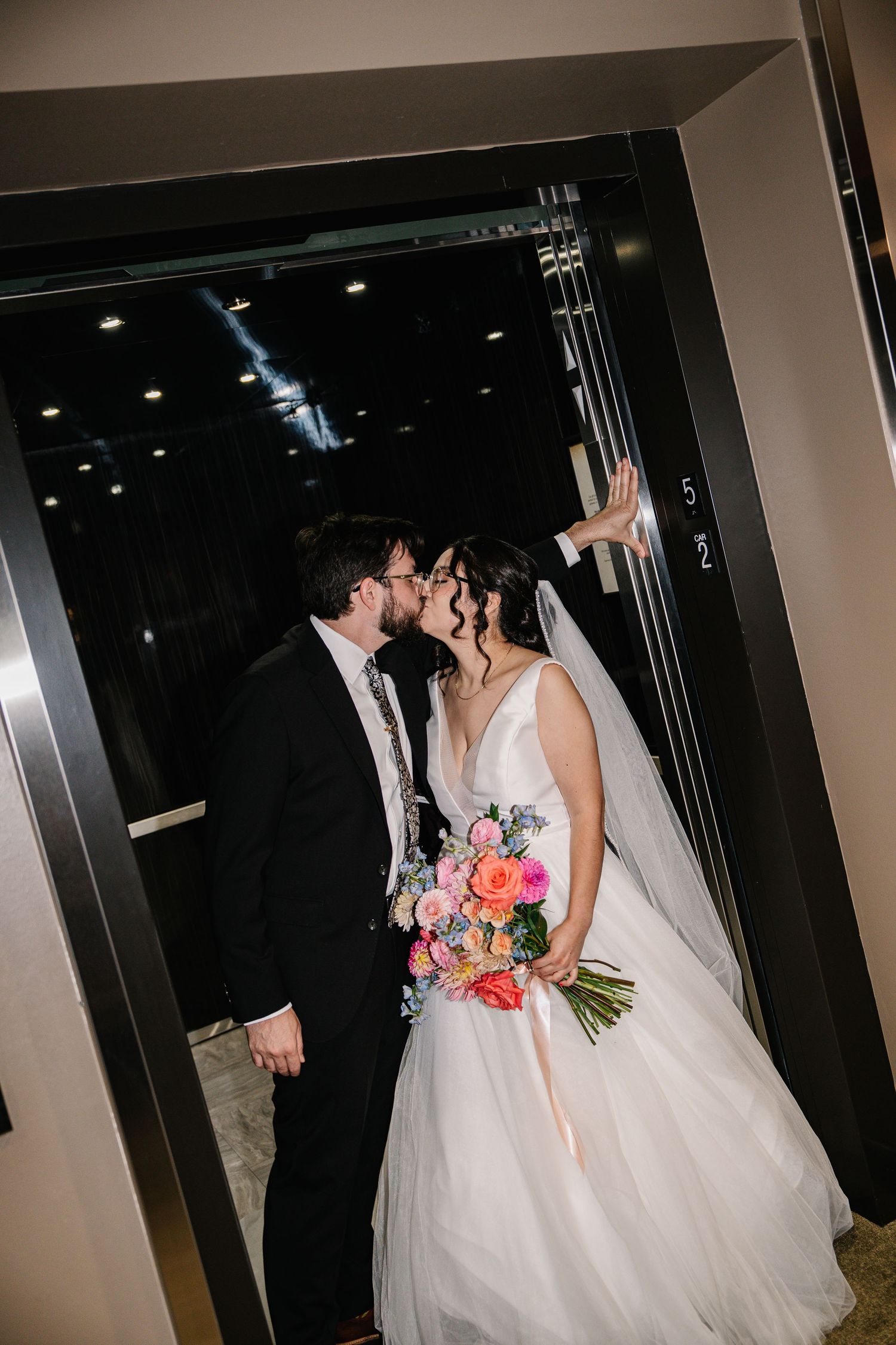 Romantic wedding couple sharing a kiss in a dimly lit hallway while holding a vibrant coral and pink bouquet.