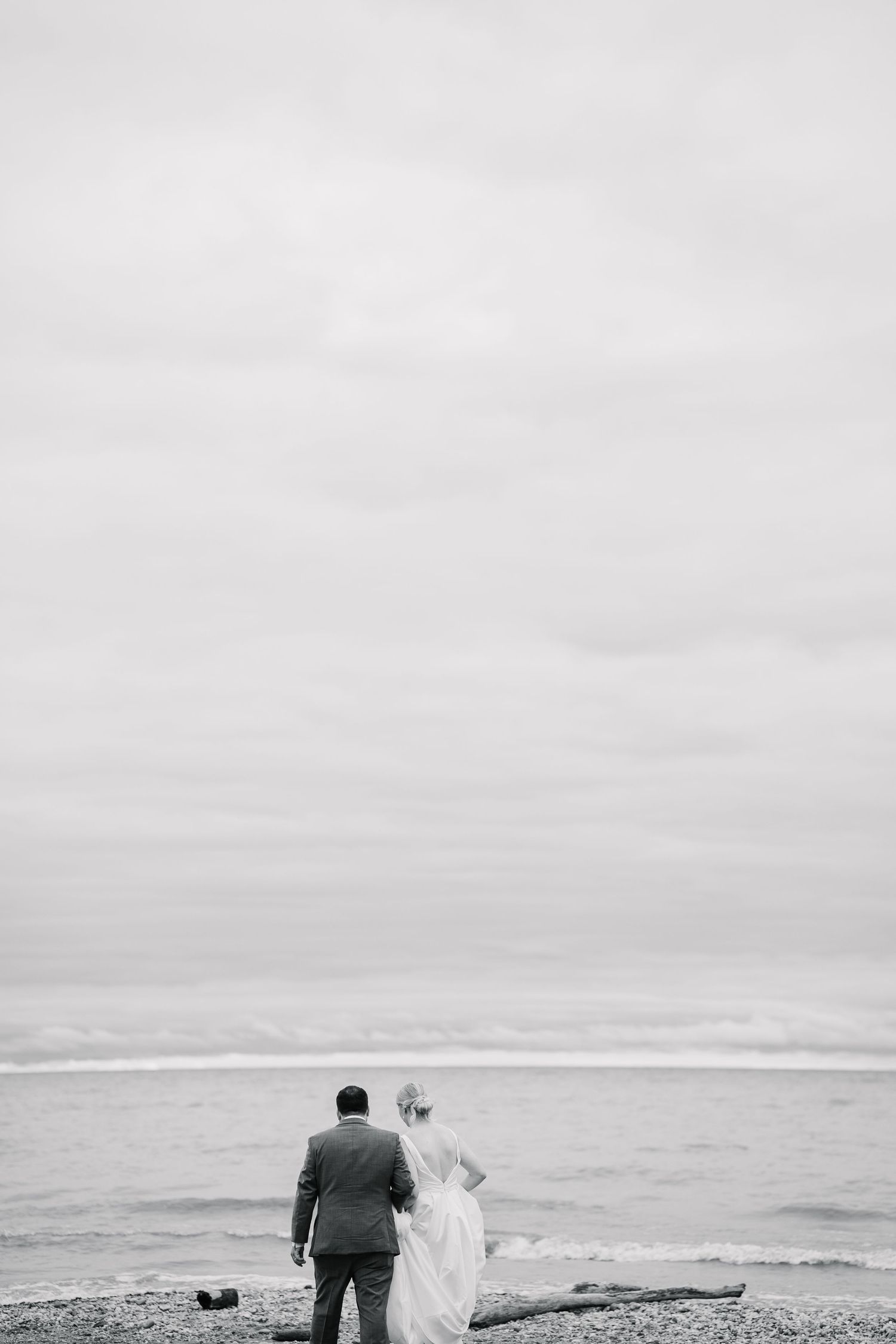 A couple in formal attire stands at the edge of a misty beach overlooking the ocean in black and white.