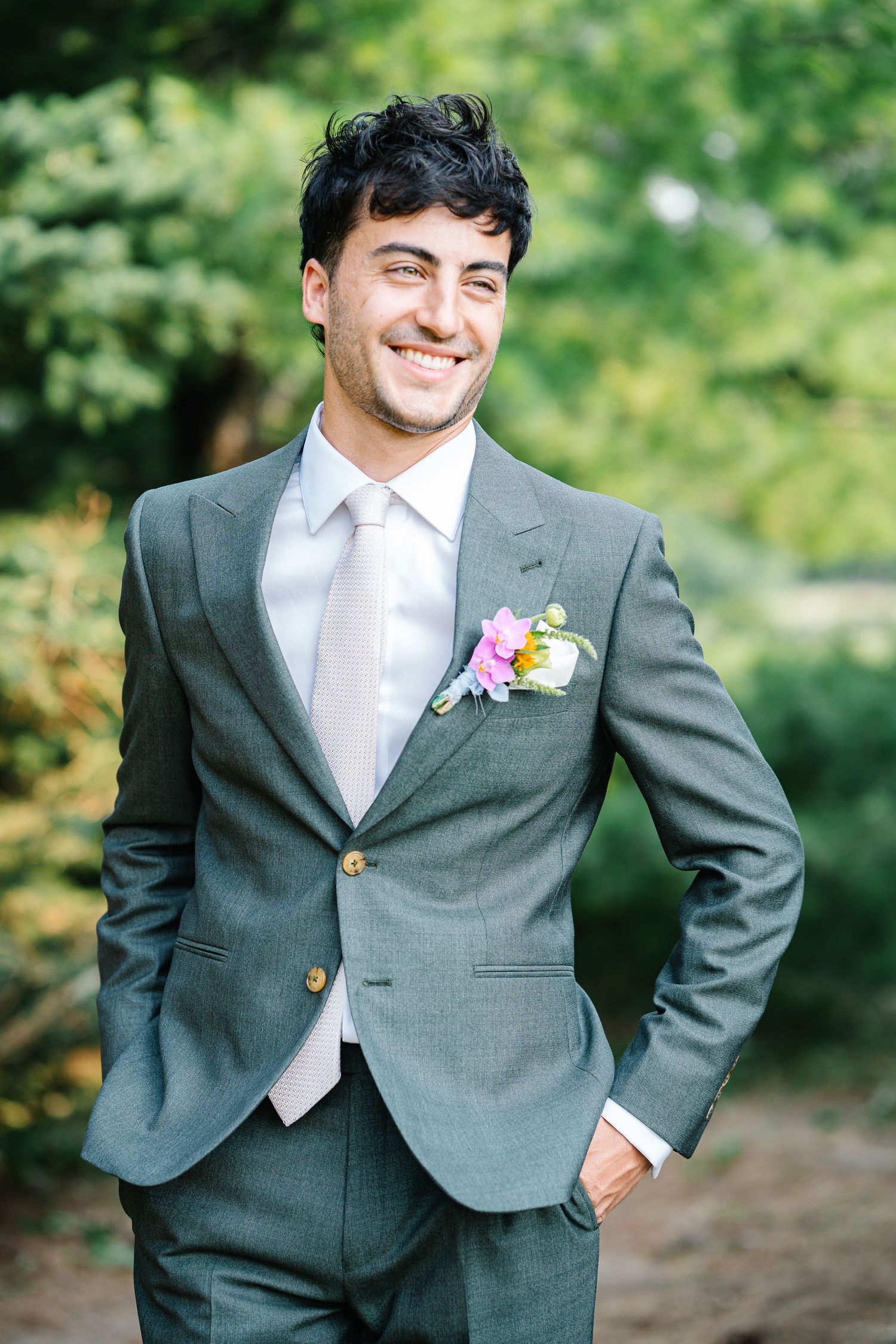 A groom in a classic grey suit with a white shirt and colorful boutonniere poses outdoors against a natural green background.