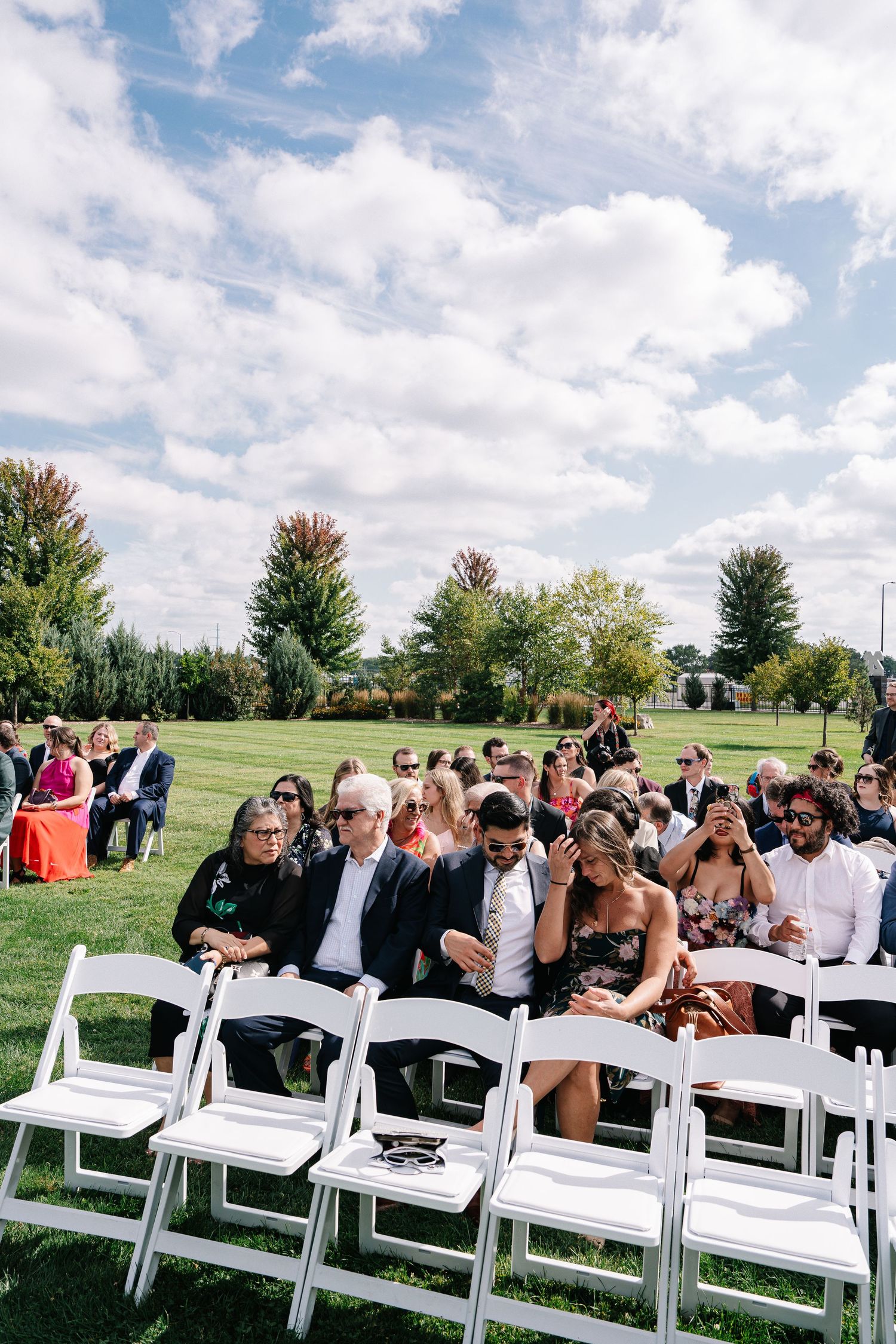 Wedding guests seated in white chairs on a sunny lawn with puffy clouds overhead.