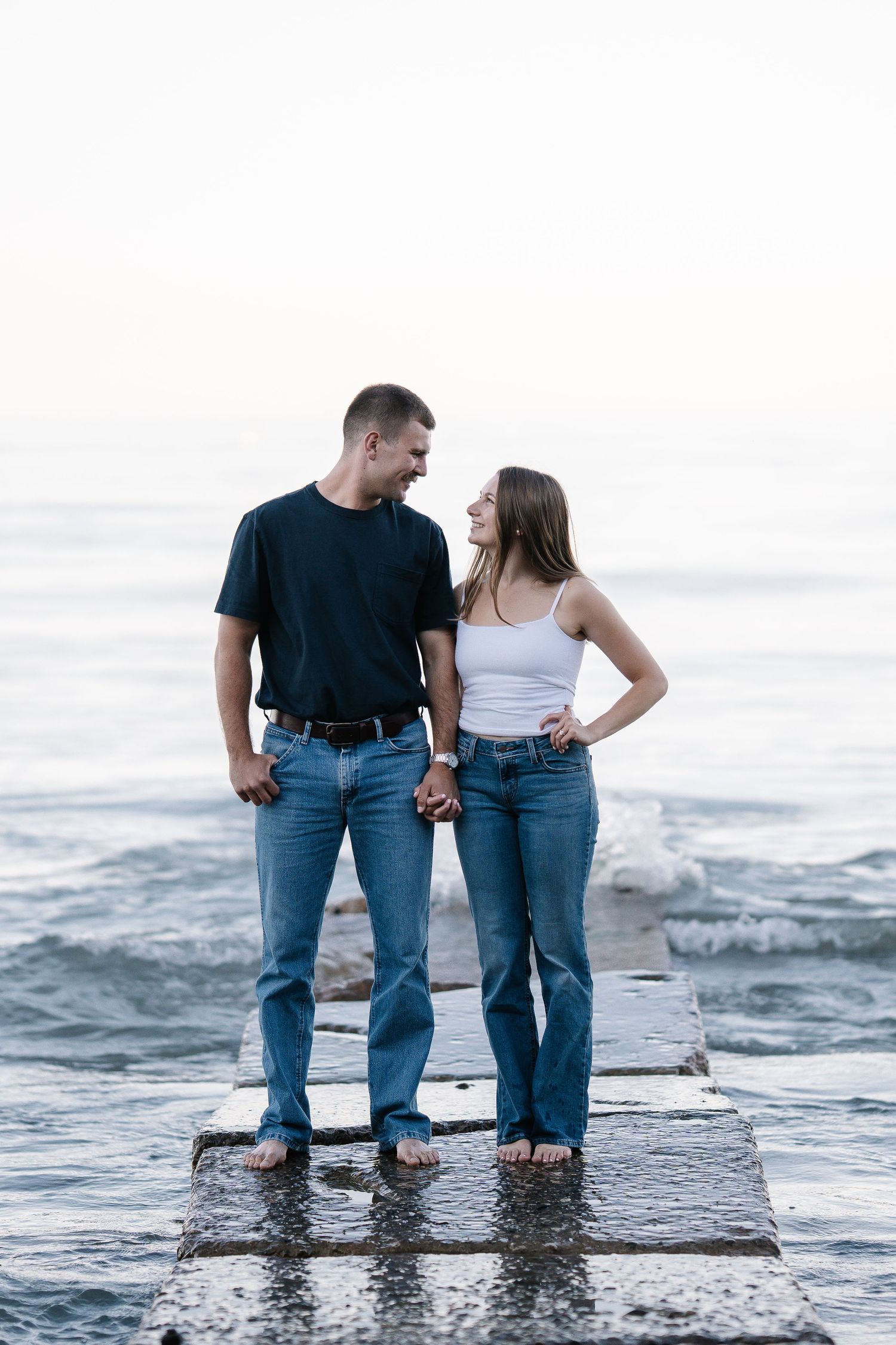 Couple holding hands on a wet pier by the ocean, wearing jeans and casual tops, smiling at each other.