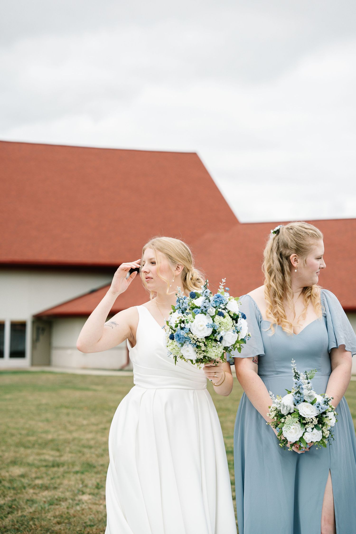 Two women in wedding attire holding blue and white floral bouquets stand together outside a barn with red roof.