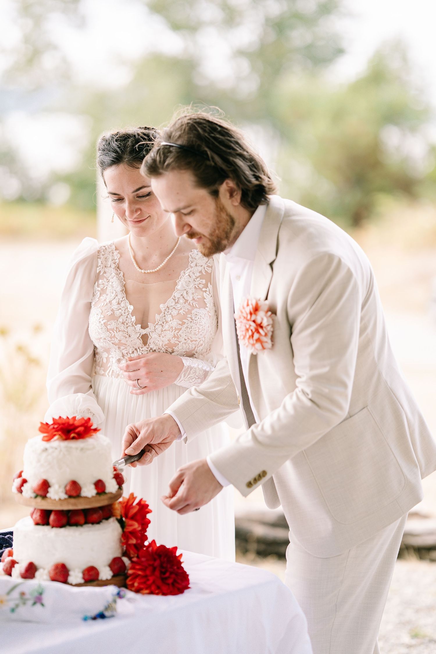 A couple cuts into their elegant white wedding cake adorned with vibrant red flowers at their outdoor reception.
