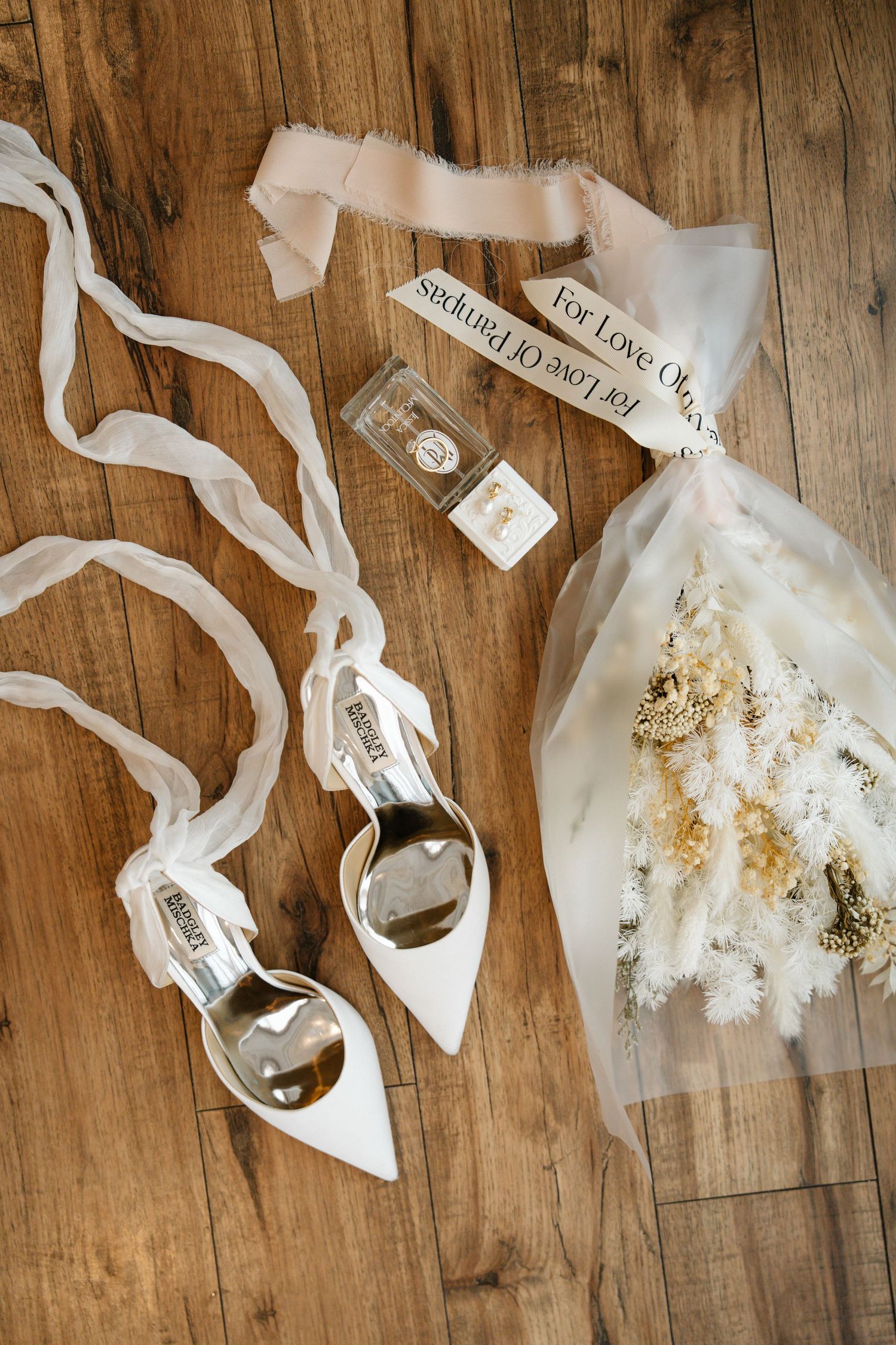 White bridal shoes, veil, and perfume bottles arranged on rustic wooden floor in wedding day flatlay.