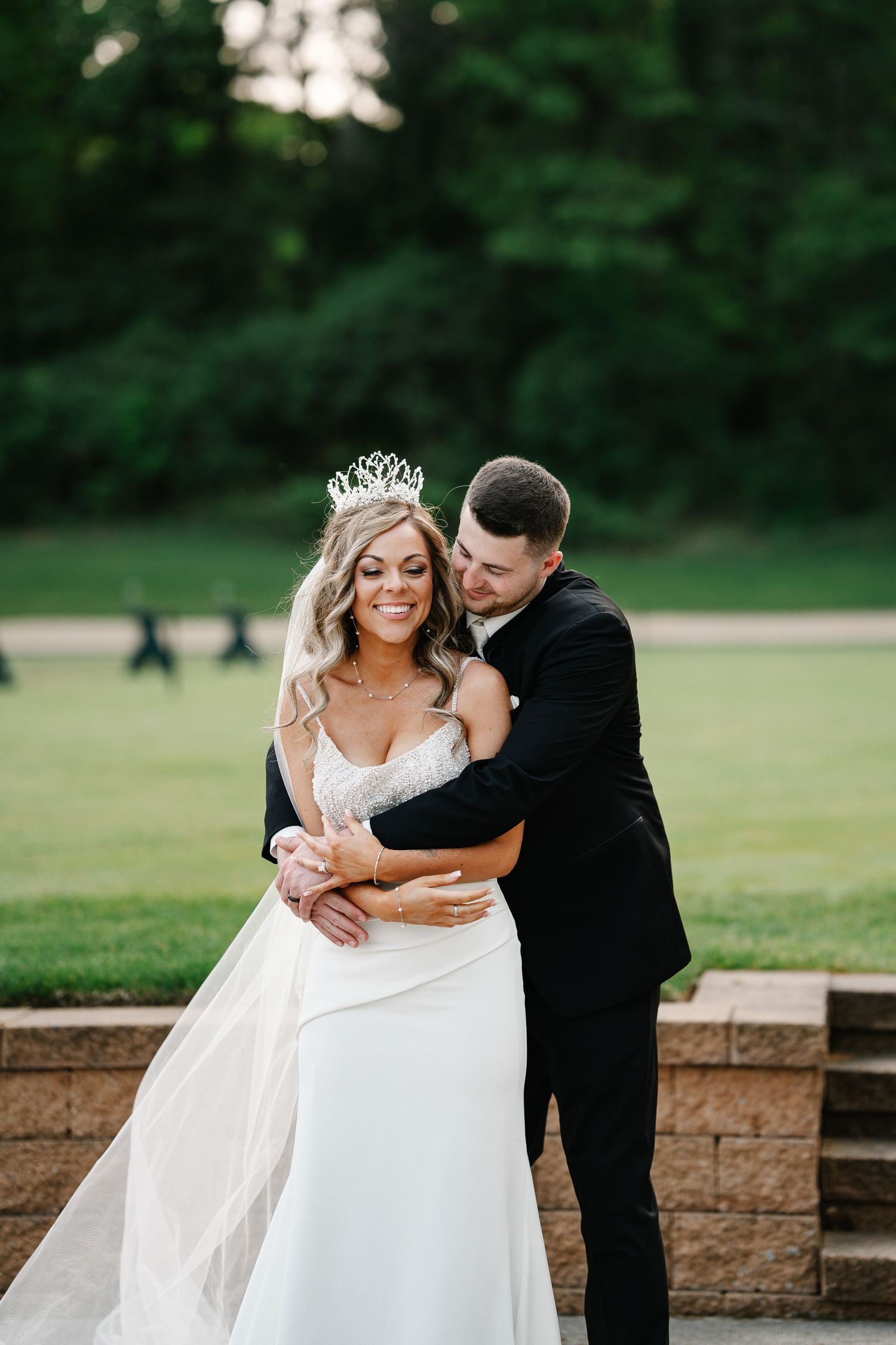 Romantic wedding couple embracing on stone wall with lush green background.