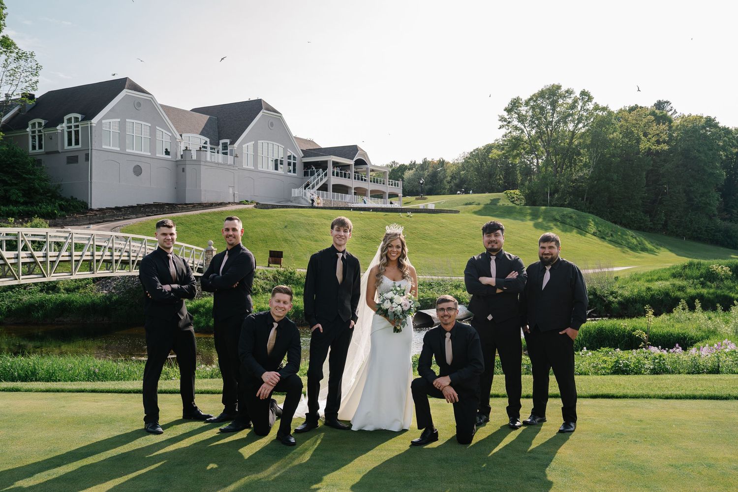 Wedding party members pose together on a lush green golf course with buildings in the background.