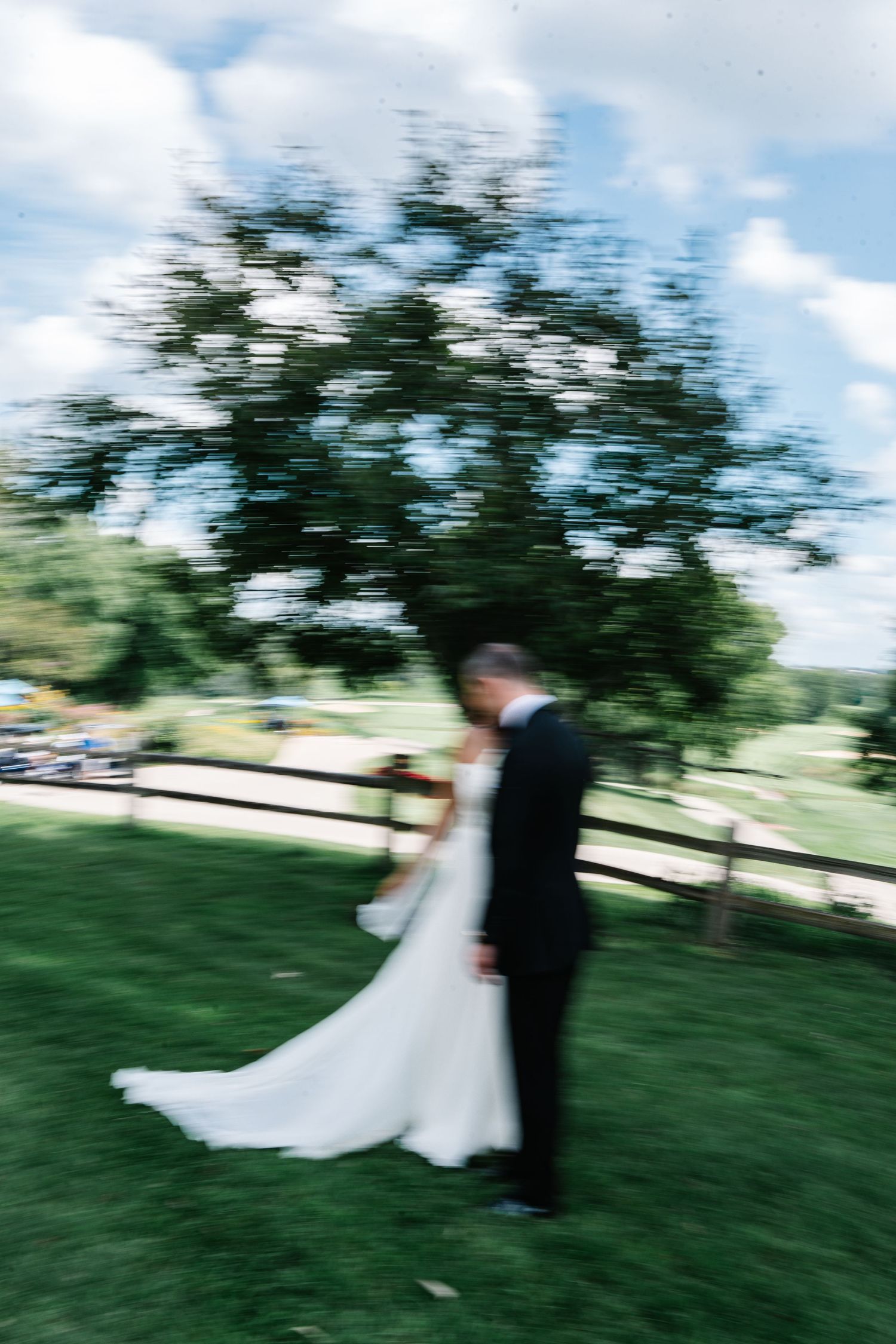 A couple in wedding attire dances together on a grassy hill beneath trees and blue skies.