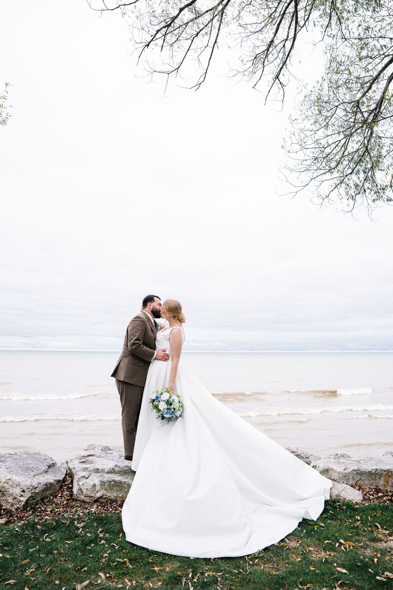 A romantic lakeside wedding scene with couple embracing on a grassy shore as waves lap at the beach.