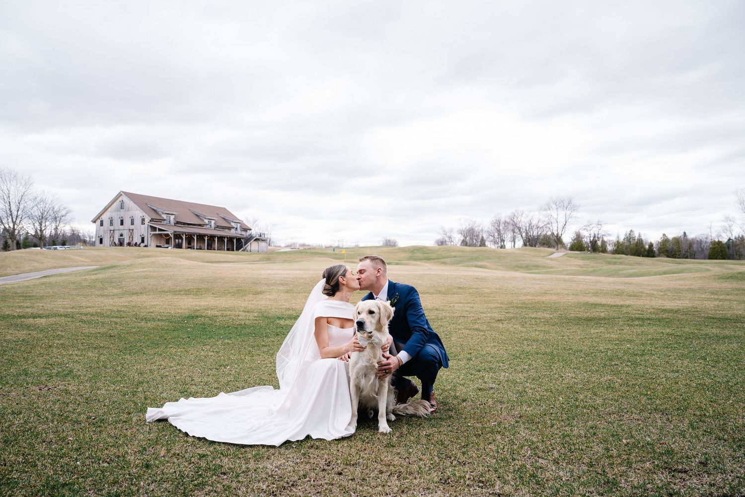 A romantic wedding couple embraces on a grassy field with a rustic farmhouse in the background.