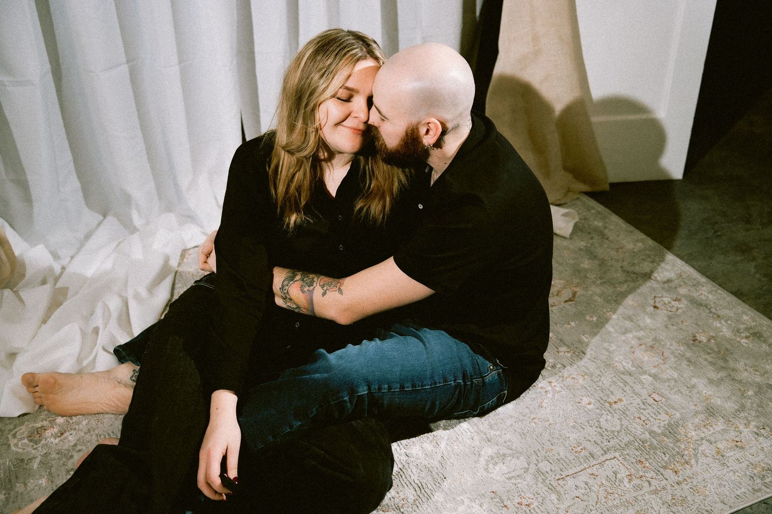 A couple in black clothing shares tender moments while sitting on a light colored floor in a dimly lit setting.