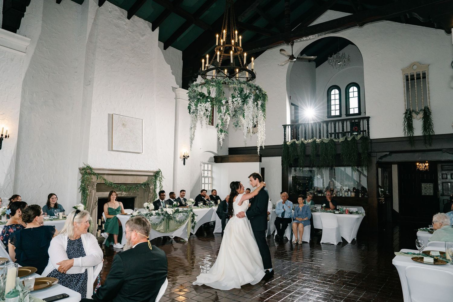 Wedding reception guests inside an elegant dark-lit ballroom with chandeliers and ornate details dancing in celebration.