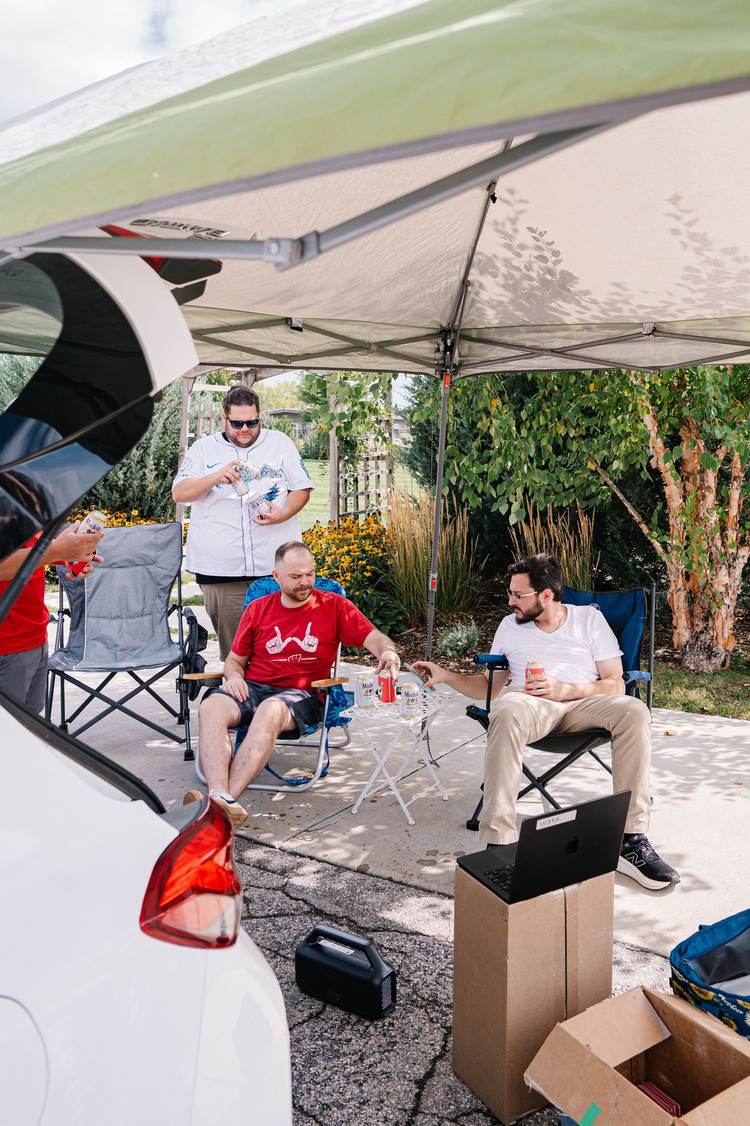 Wedding party members relax in camping chairs under a white canopy tent during outdoor preparations.