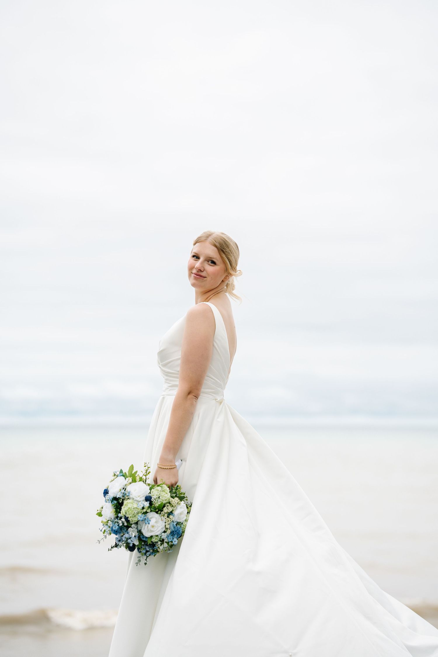 A bride in a flowing white wedding dress holds a blue and white bouquet while standing on a beach against an overcast sky.