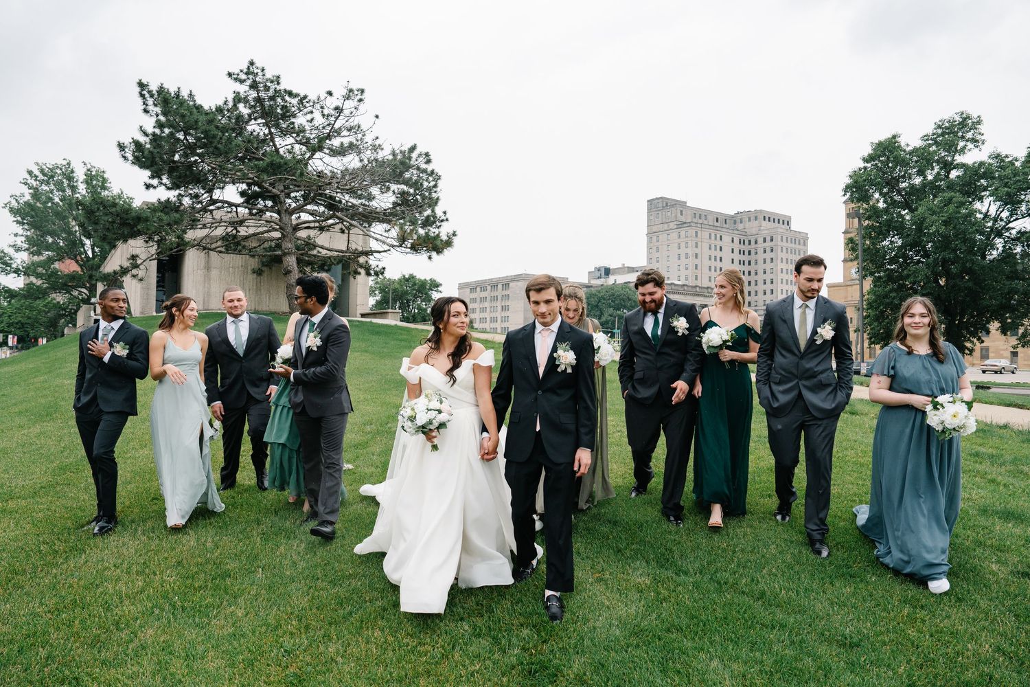 Wedding party walks together on grassy lawn with modern building in background.