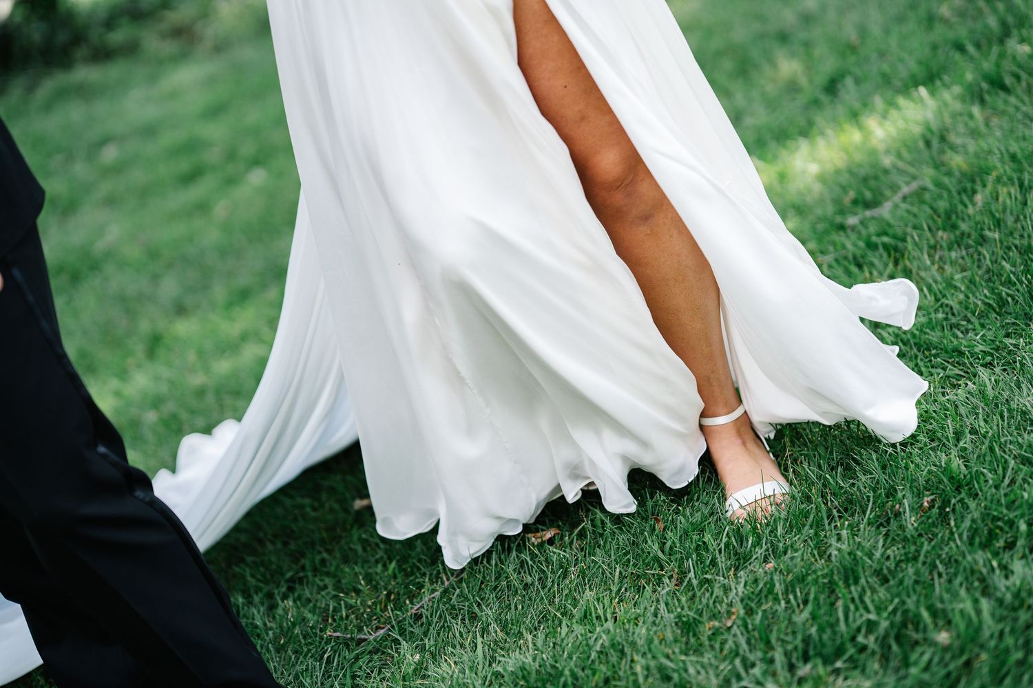 A bride's white flowing dress and bare feet are captured in motion across lush green grass.