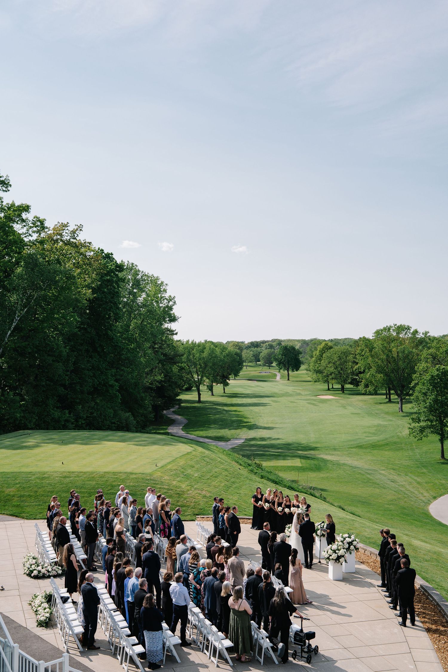 Outdoor wedding ceremony setup at a golf course with guests gathered on a stone patio.