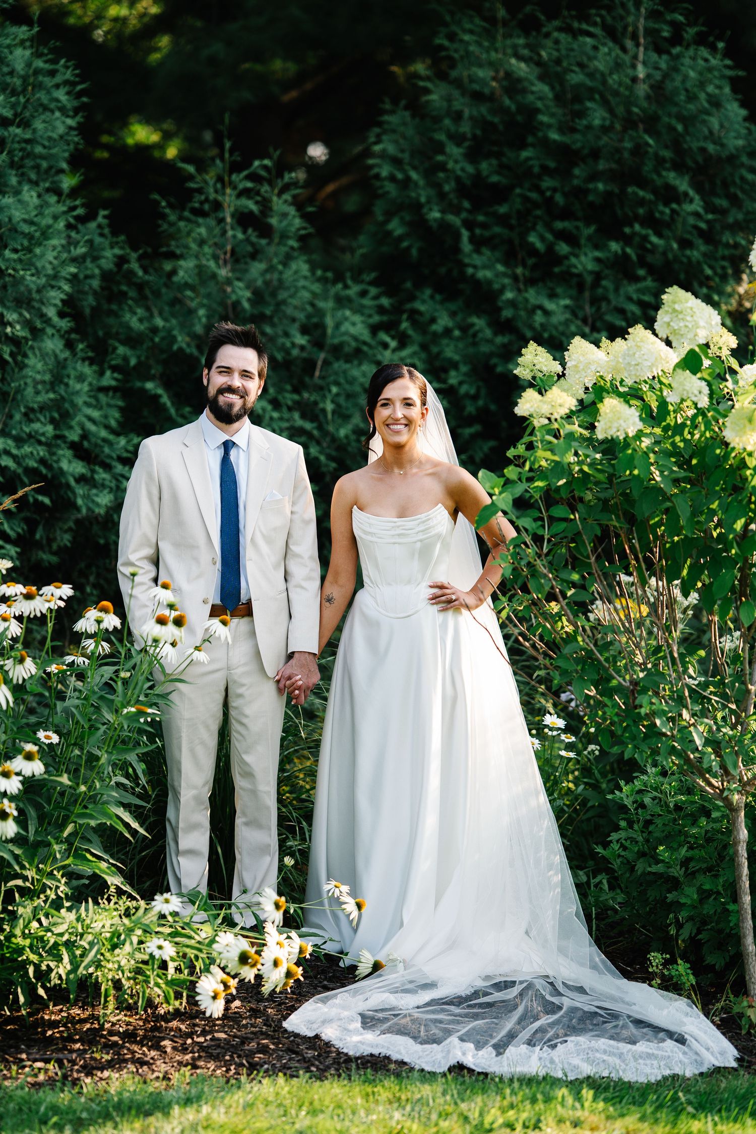 A couple stands together in a lush garden setting with hydrangeas and greenery on their wedding day.