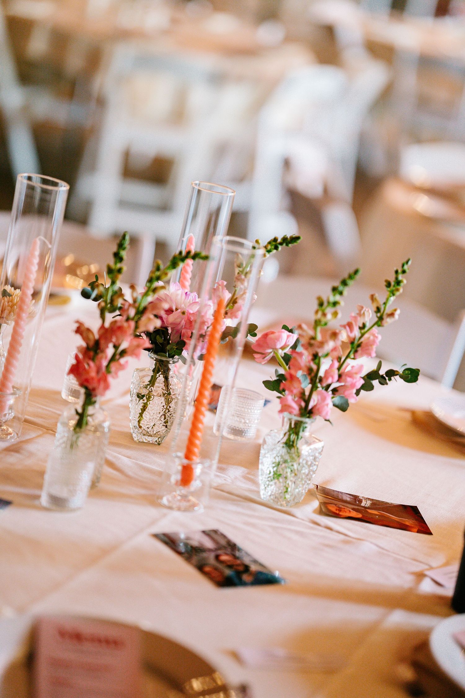 Small pink flowers in glass vases line up along a white tablecloth at an elegant wedding reception table setting.