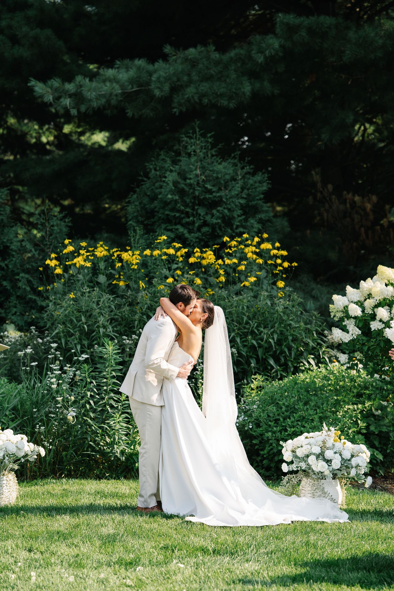 A romantic wedding couple embraces in a lush garden setting with yellow and white flowers blooming around them.