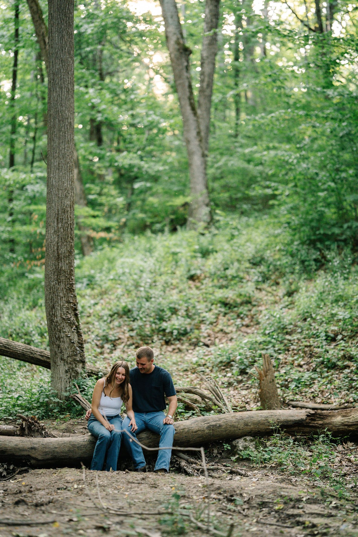 Couple sitting together on a fallen log in a lush green forest, sharing a quiet moment outdoors.