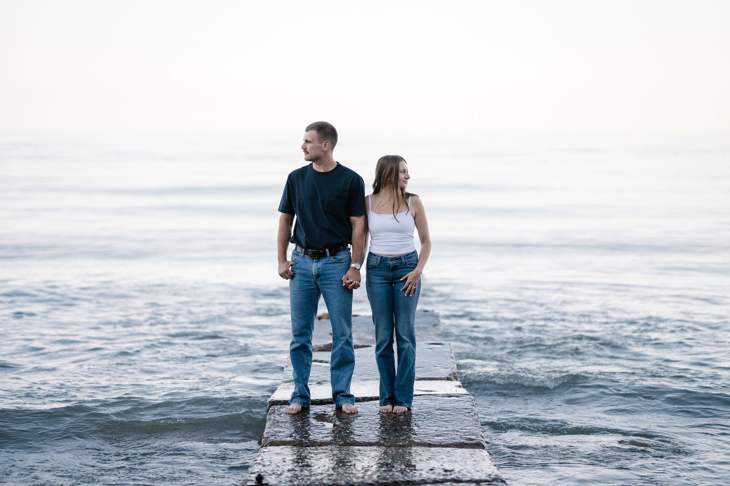 Couple holding hands standing on a dock overlooking a calm ocean horizon on a clear day.