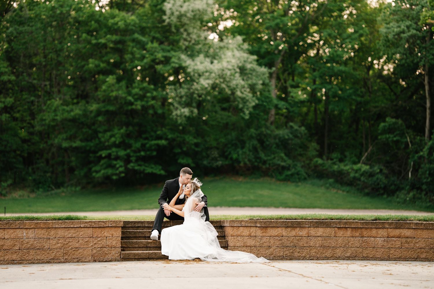 Scenic outdoor wedding photos at sunset showing couple in formal attire posing on stone wall amidst lush greenery.
