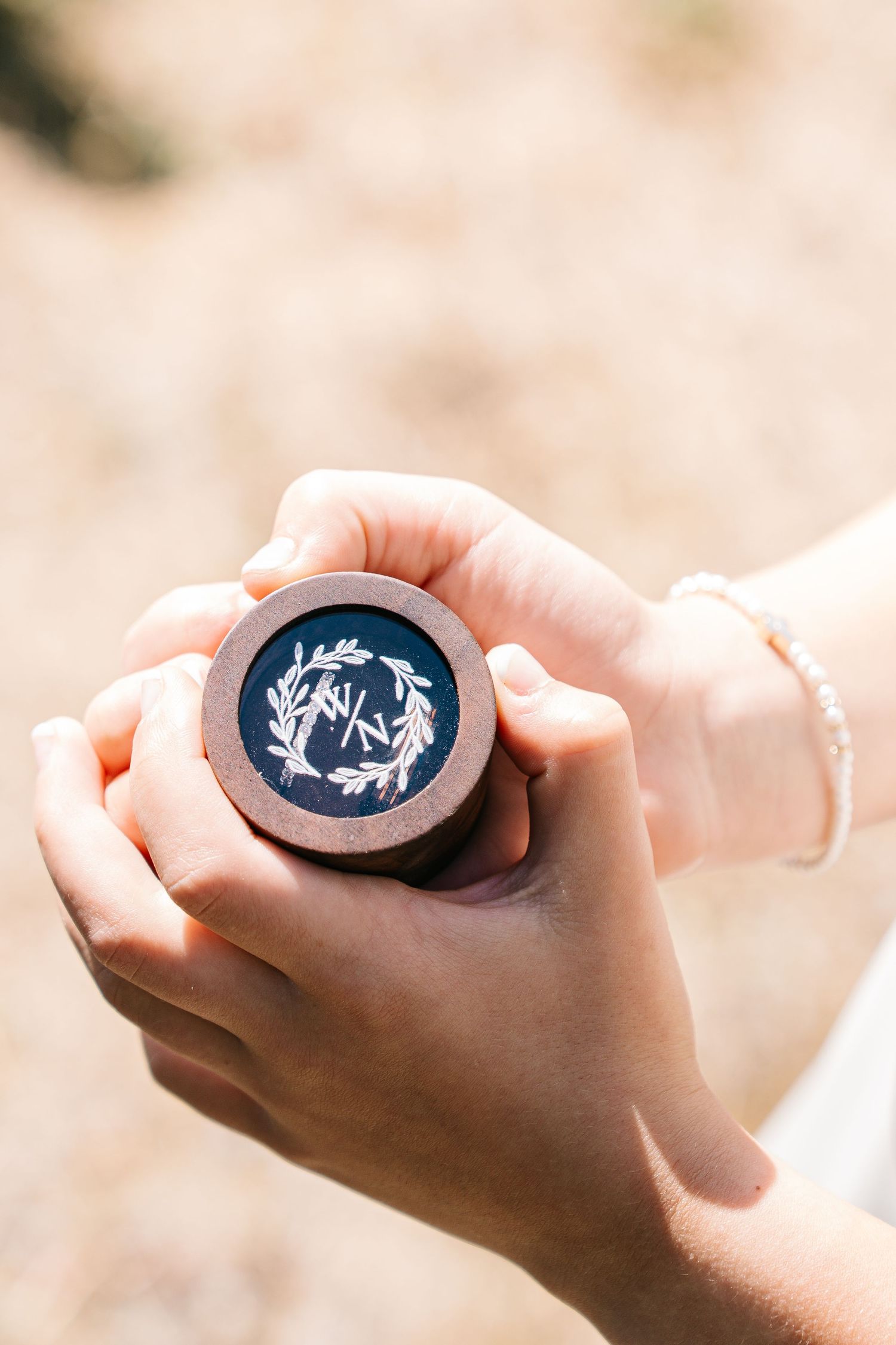 Hands holding a small wooden ring box with a monogrammed design featuring a laurel wreath.
