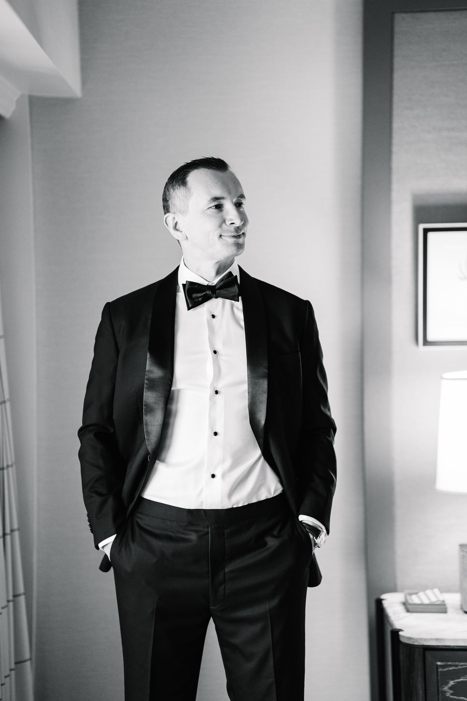 Black and white photograph of groomsmen in formal tuxedos standing in a bright room before a wedding ceremony.